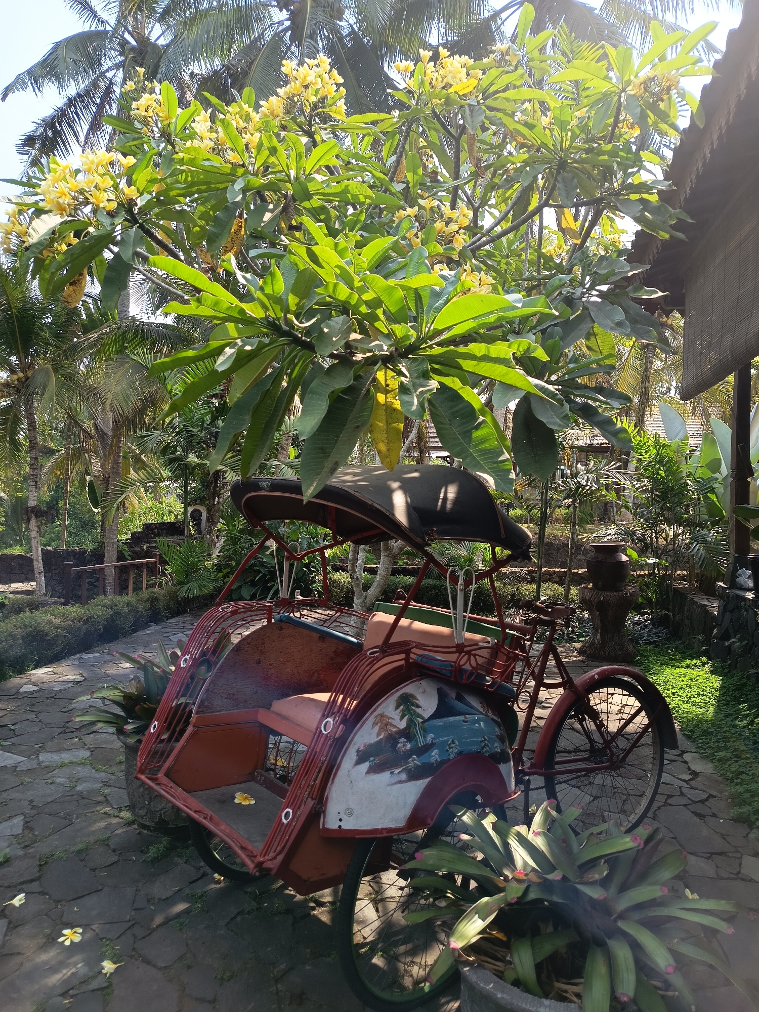 The image captures a bright, sunny outdoor scene in Ngaglik, Indonesia, featuring a traditional red pedicab parked on a stone pathway. The pedicab has a canopy and is adorned with a painted mural depicting palm trees and a coastal landscape on its rear side. Surrounding the pedicab is lush tropical vegetation, including palm trees and a flowering tree with yellow blossoms overhead. In the background, traditional Indonesian architecture with thatched roofs is visible, along with more dense foliage. The ground is paved with irregular stone slabs, and a few yellow flowers are scattered on the pathway. The scene conveys a sense of a tranquil, tropical environment.