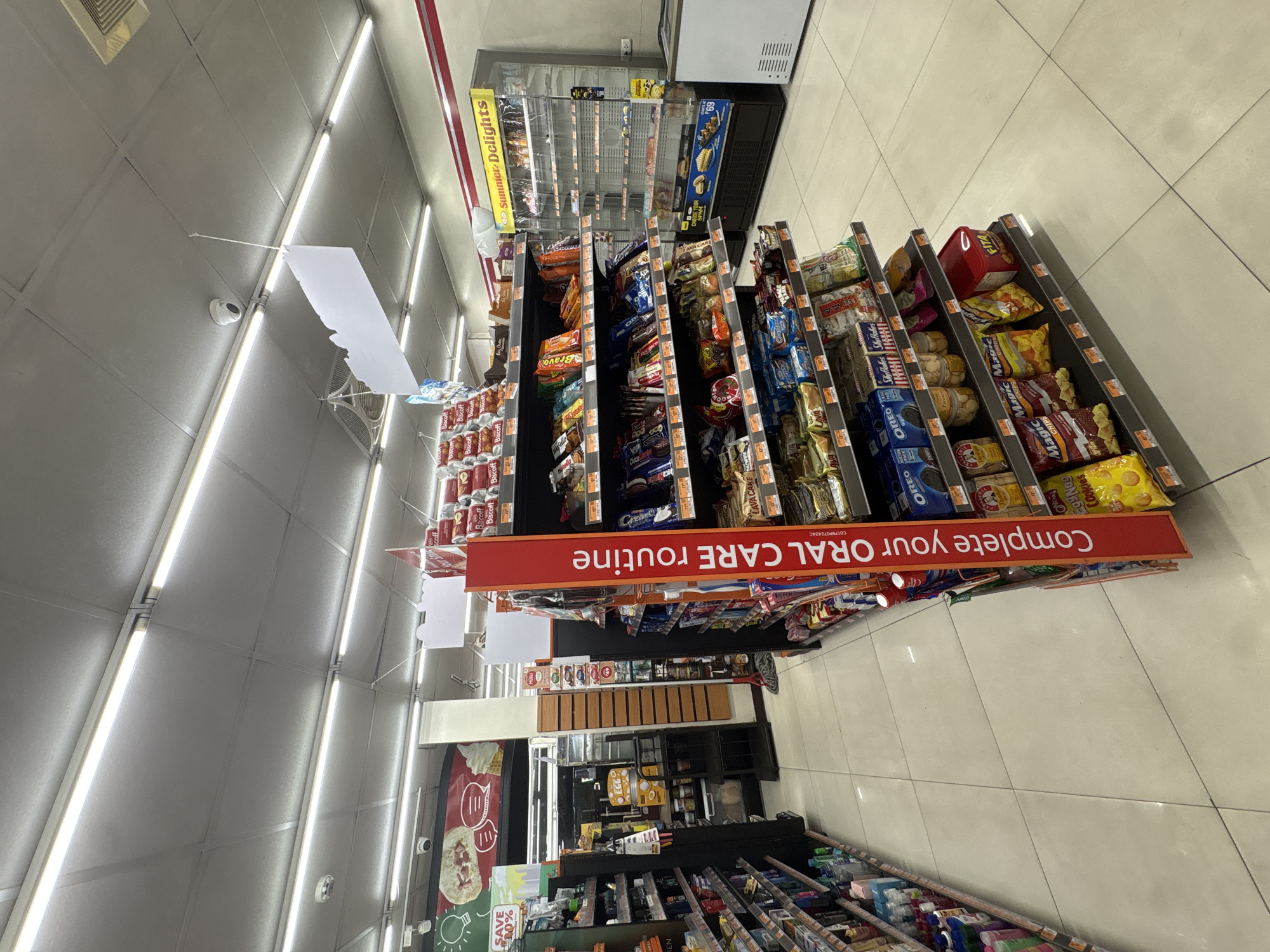 This image depicts the interior of a convenience store located in Davao City, Philippines. The store features a white drop ceiling with multiple linear fluorescent light fixtures and visible dome-shaped CCTV cameras. The floor is composed of light-colored square tiles with dark grout lines.
In the foreground and mid-ground, multiple black metal shelving units are arranged, forming aisles. A prominent red vertical sign, bearing the white text "Complete your ORAL CARE routine," is attached to a shelving unit on the right side of the primary aisle. This unit displays various packaged snack items, including Biscoff biscuits, Oreo cookies, Lava Cake, Magic Chips, and Egg Nogg Cookies. A shelving unit on the left side of the main aisle is stocked with personal care products and toiletries.
In the background on the right, a large glass display case with yellow "Summer Delights" signage is visible; it appears mostly unstocked. Below this, a smaller display promotes "CHOOSE YOUR SPAM" items, with pricing starting at "₱69." A white chest freezer is positioned next to this display. On the upper left wall, a red banner features images of food items and text, including "SAVE 10%." A black utility cart and a mop are discernible in a secondary aisle in the left background. No individuals are present in the depicted scene.