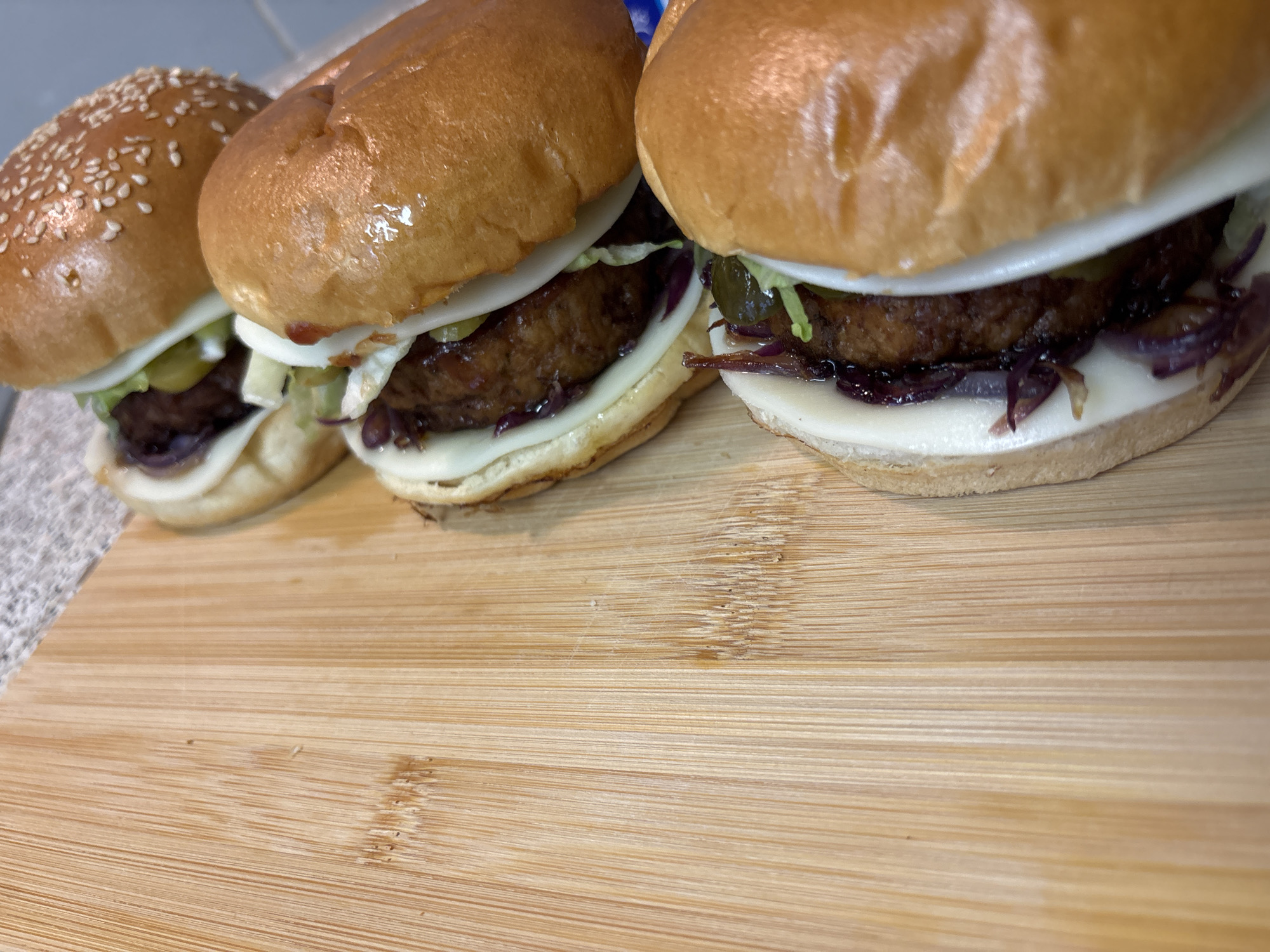 Three small burgers, possibly sliders, are arranged in a diagonal line on a wooden cutting board. The burgers are topped with sesame seeds on their top buns, with one burger showing a full view of its sesame seed topping. The burgers appear to be composed of a patty, cheese, lettuce, sliced red onion, and pickles. The cutting board has a visible grain pattern. The background is out of focus, showing hints of a countertop and a bluish surface. The image was taken from a high-angle perspective, looking down at the burgers.