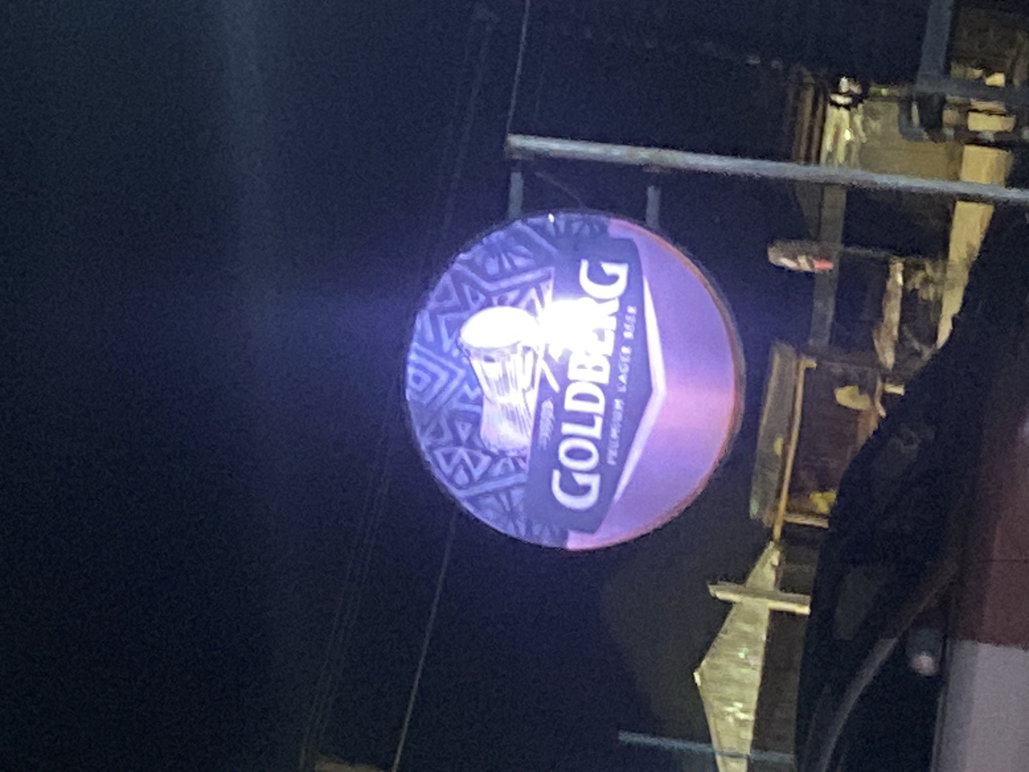 The image shows an illuminated Goldberg beer sign at night. The sign is round, with a blue patterned background featuring a beer mug graphic and the "Goldberg" logo in white, overlaid with a purple hue. The sign is mounted on a metal structure, possibly a pole or building support. In the background, parts of a building or structure are visible, along with electrical wires. The surrounding environment appears dark, indicating nighttime. The location is likely Jos, Nigeria, based on the product advertised.