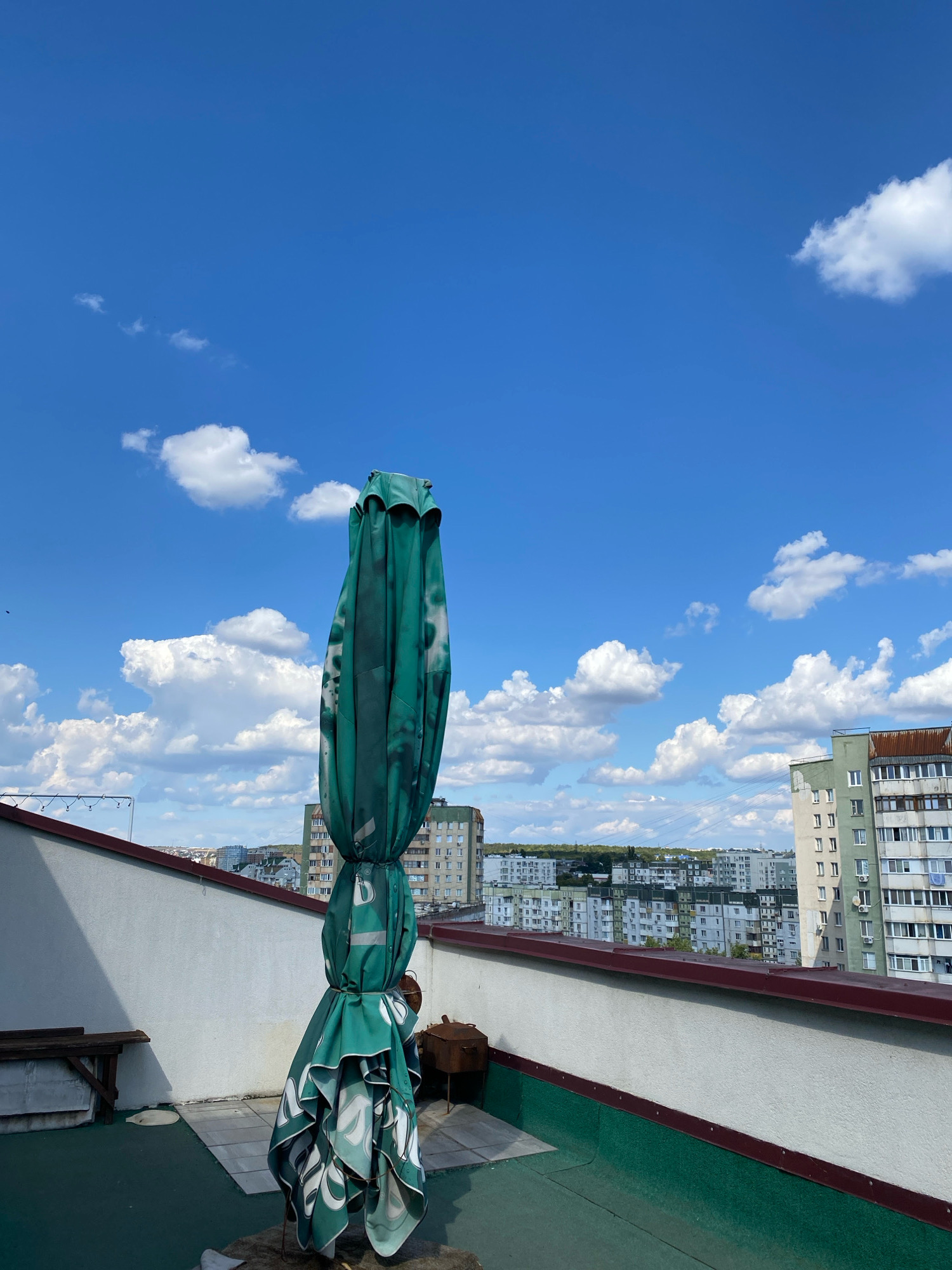 This is a medium-shot, eye-level, vertical, exterior photograph of a closed green umbrella on a rooftop in Chișinău, Moldova. The umbrella is in the foreground, on the right side of the frame, and takes up a significant portion of the image. It is tied at the top and bunched up towards the bottom, with a white and green patterned fabric visible at the base. To the left of the umbrella is a wooden bench, and behind it is a white wall. The rooftop surface is a dark green color. In the background, across the skyline, are numerous Soviet-era apartment buildings, predominantly grey and white, with some showing signs of wear and tear, like rust on the roofs. The sky is a vibrant blue with scattered white, puffy cumulus clouds. The overall impression is a daytime scene, likely late spring or summer, with fair weather.