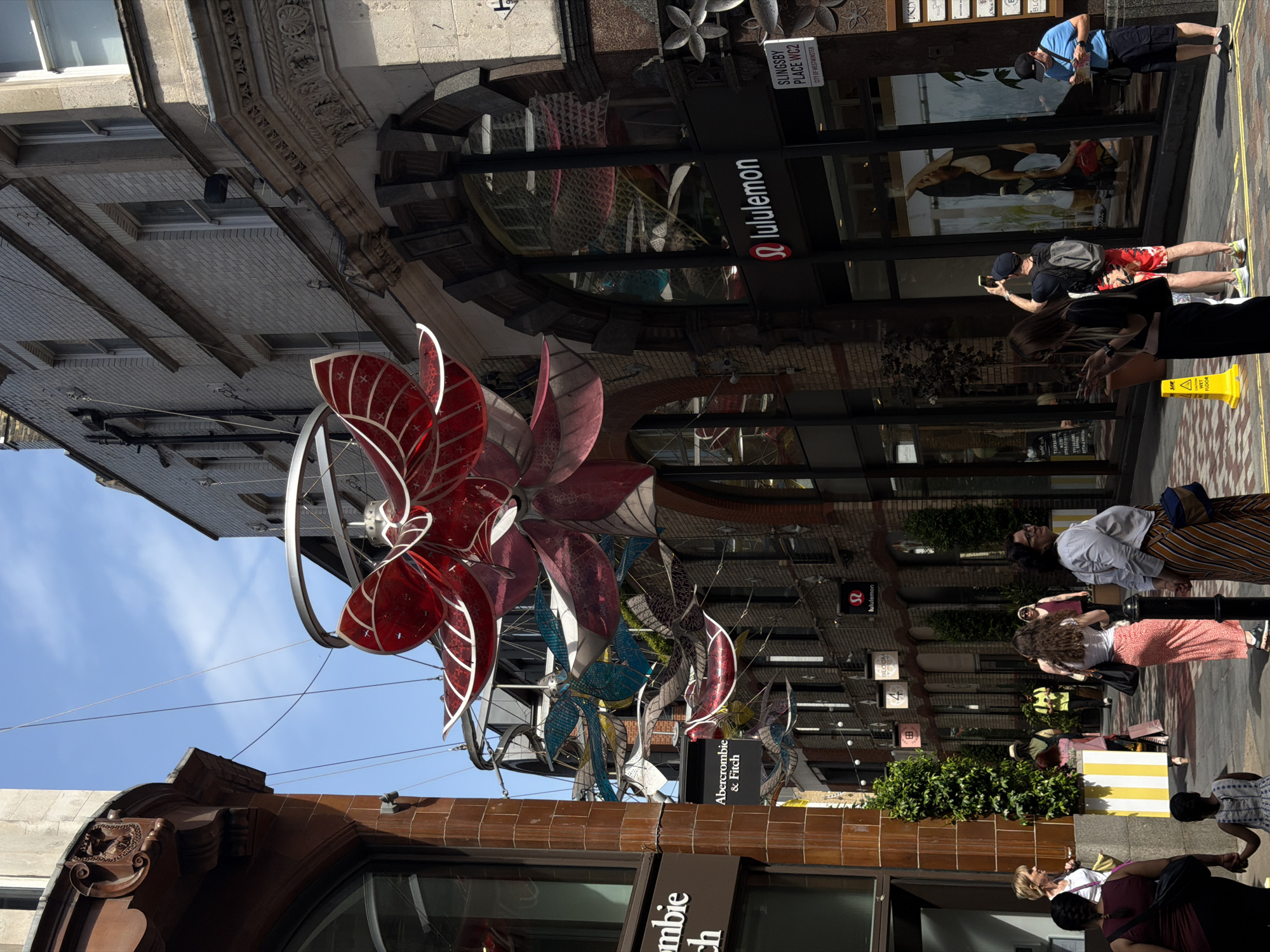 The image captures a street scene in London, United Kingdom, with a focus on storefronts and pedestrians. The street is adorned with overhead floral decorations, predominantly red, with some blue and white accents. Below these decorations are the storefronts of several businesses, including Abercrombie & Fitch, and Lululemon, the latter with the street name "Slingsby Place WC2" and "City of Westminster" marked on a sign.
Pedestrians are present on the pavement, some appearing to be shopping. Several individuals are visible taking photographs with their mobile phones, suggesting the floral installations are a point of interest. The perspective is a street-level view, looking upwards towards the storefronts and floral decorations. The sky is visible in the upper left corner.