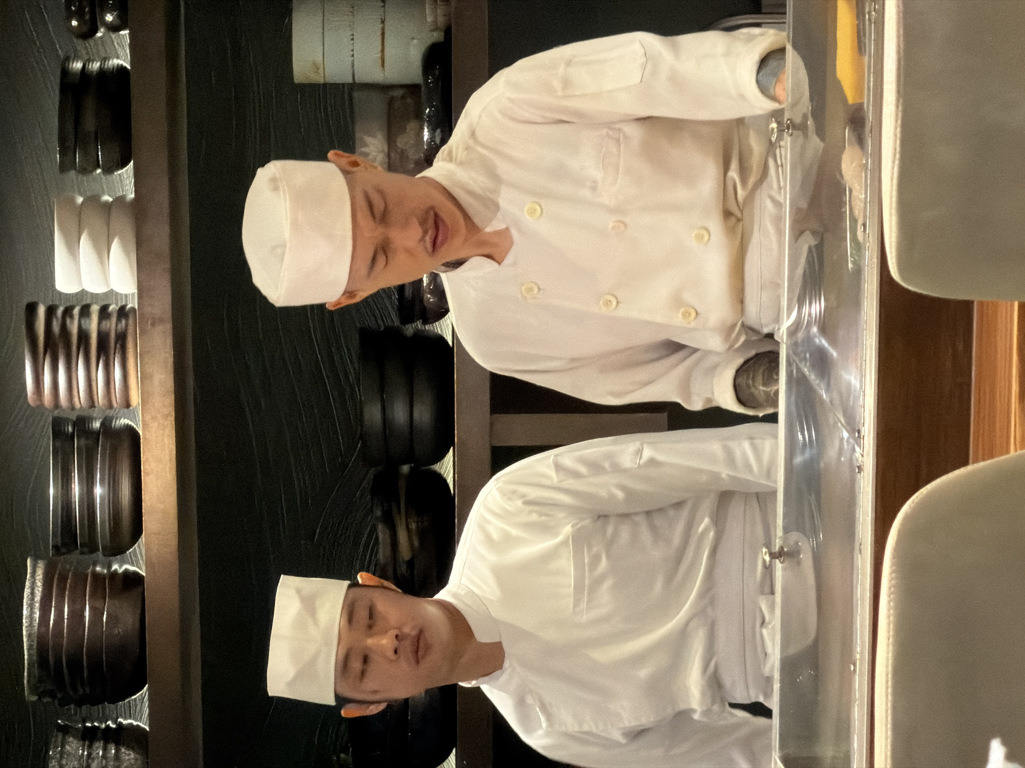 In San Sai Luang, Thailand, two Asian chefs wearing white chef coats and hats stand on opposite sides of a transparent barrier, likely in a restaurant kitchen. The upper chef's face displays a slight grimace, while the lower chef maintains a neutral expression. A wall to the left holds arranged kitchenware: dark bowls and utensils in a vertical row. Behind the chefs, shelves hold stacks of black plates. A chair and wooden paneling are visible on the right.