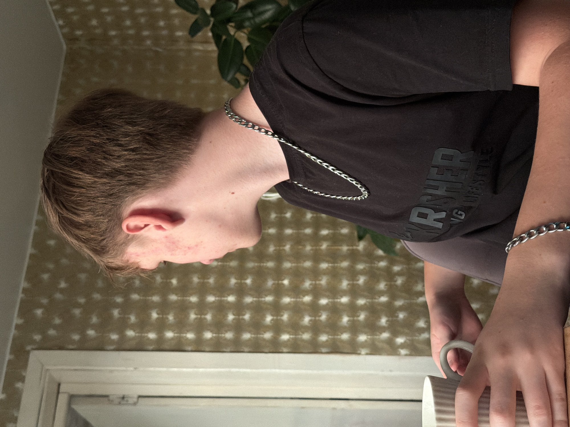 A young male with light brown hair and fair skin is seen from the side, looking out a window. He is wearing a black t-shirt with a logo that appears to read "THRASHER" and "LIFESTYLE" in grey text. He has a silver chain necklace and a silver bracelet on his left wrist. His left hand is holding a white mug with a ribbed texture. The background features a wall with a textured, light brown wallpaper and a hint of greenery from a plant to the right. A white-framed window is visible on the left. The lighting suggests an indoor setting, possibly during the daytime. No specific location in Mezhevoy, Russia, can be determined from the visual information.