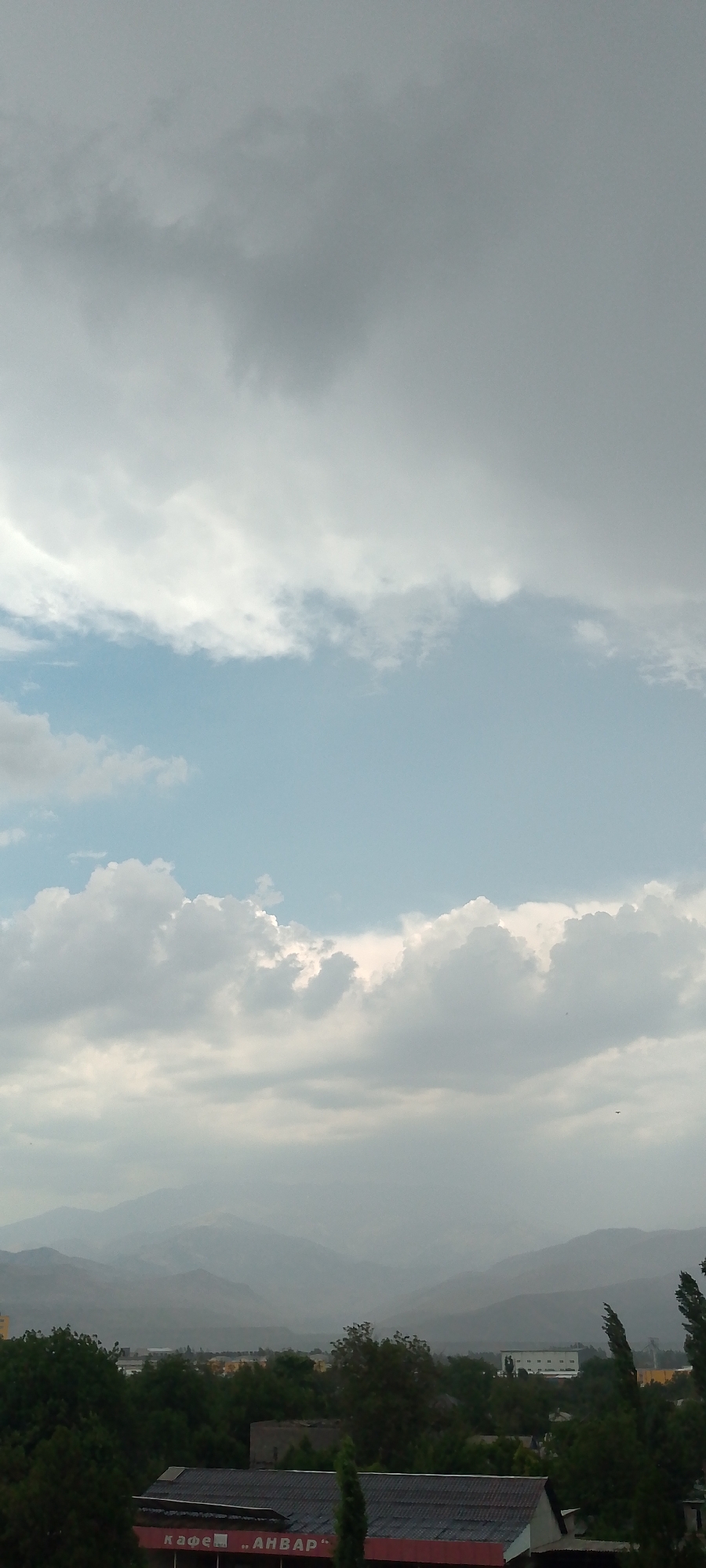 The image depicts a landscape with a vast sky filled with clouds dominating the upper portion. The clouds are a mix of dark gray and white, suggesting possible impending rain or overcast conditions. Below the clouds, a light blue sky is visible in some areas. In the distance, a range of hazy mountains can be seen, their shapes blurred by atmospheric conditions. The middle ground of the image features a town or city with buildings and dense green trees. A red and gray building with a sign that reads "КАФЕ ,,АНВАР"" in Russian is prominently visible in the foreground. The overall impression is one of a somewhat overcast day in a semi-urban or rural setting with mountainous terrain in the background. The location is identified as Angren, Uzbekistan, which is known for its industrial areas and surrounding natural landscapes.