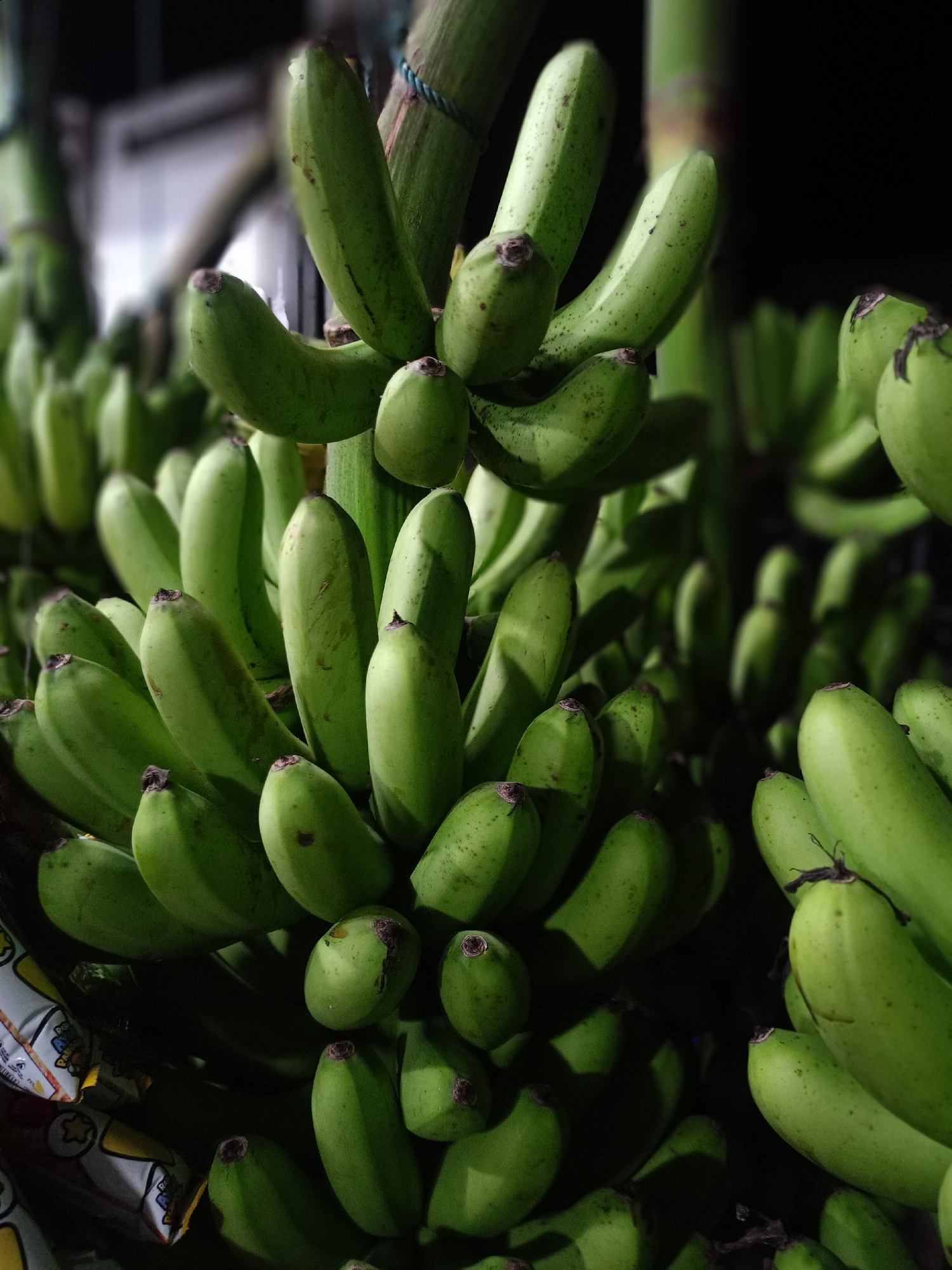 The image shows several bunches of unripe green bananas, possibly in a market setting or storage area in Uteunreutoh, Indonesia. The bananas appear to be hanging or stacked, with some partially obscured by others. The lighting is somewhat dim, casting shadows across the bunches. A glimpse of a product packaging is visible in the lower left corner, adding to the sense of being in a commercial environment.