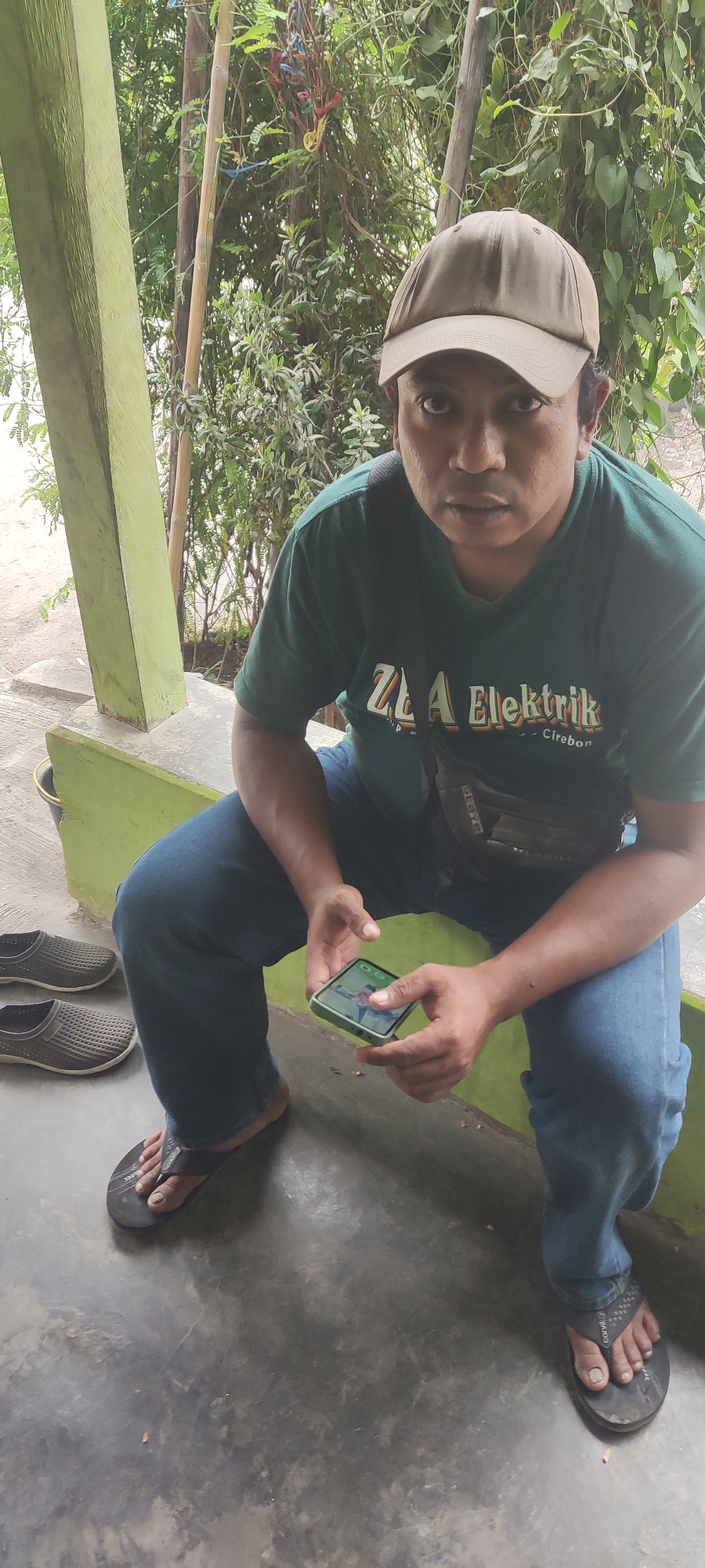 A man wearing a green t-shirt and blue jeans sits on a green bench outdoors, holding a smartphone. He has a light brown baseball cap on his head and a dark bag slung across his chest. In the background, lush green foliage and bamboo stalks are visible. Two pairs of grey slip-on shoes rest on the concrete floor to his left. The scene appears to be in Karangwangun, Indonesia.