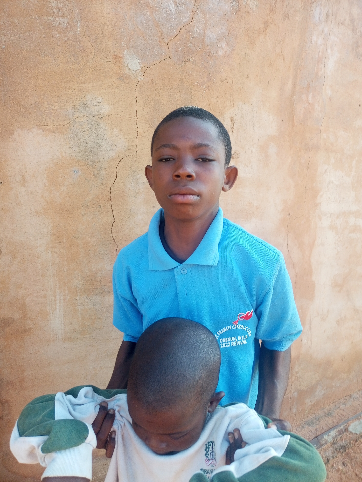 Two young Black boys are pictured in front of a tan, textured, cracked stucco wall. The boy in the foreground is facing away from the camera, with his head bowed, wearing a light-colored shirt with green and white striped cuffs and sleeves. The boy in the background stands behind him, looking directly at the camera. He has short, dark hair and is wearing a bright blue polo shirt with a crest on the left side that reads "ST. FRANCIS CATHOLIC CC" and "OREGUN, IKEJA 2022 REVIVAL". His arms are around the boy in the foreground. The image appears to have been taken outdoors during the day. The setting is a wall in Tempure, Nigeria.