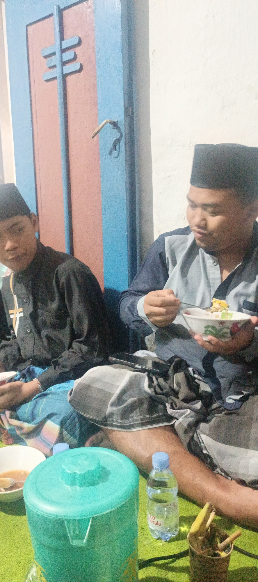 The image shows two young men wearing traditional Muslim attire, including peci caps and sarongs, sitting on a green carpet. The man on the right is eating from a bowl with a spoon. In the foreground, there is a large teal colored water dispenser and a bottle of "Le Minerale" water. A small cup filled with what appears to be cigarettes or thin sticks is also visible. Behind the men is a wooden door painted blue and salmon pink, with a handle and a key. The wall next to the door is white. The lighting suggests it is indoors and likely evening or night. The scene appears to be casual and domestic.