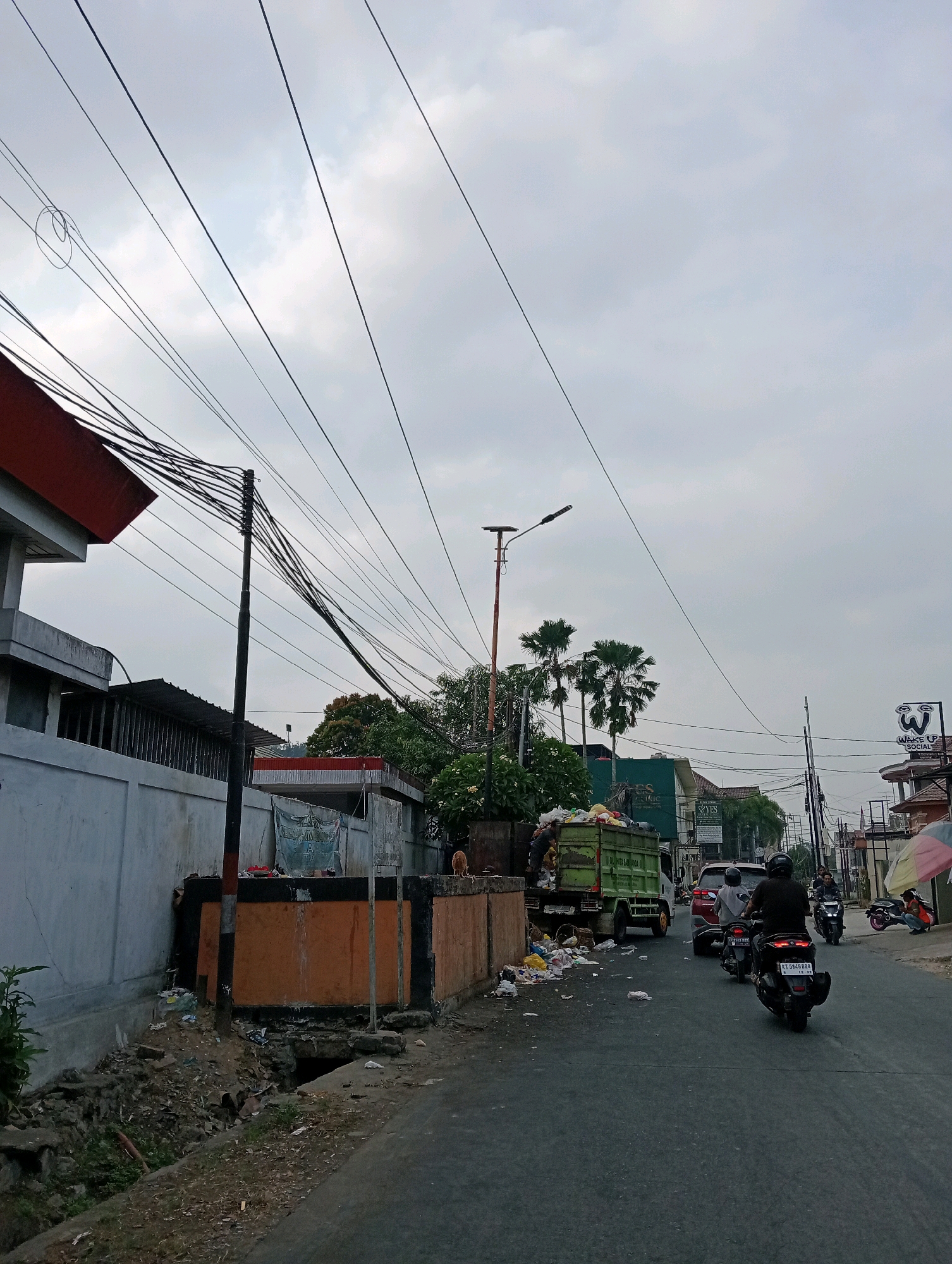This image depicts a street scene in Samarinda, Indonesia, on a cloudy day. The foreground shows an asphalt road with several motorcycles and a car. A green garbage truck is parked on the left side of the road, partially obscured by debris and trash scattered around it. A person appears to be working on or near the truck.
To the left of the road, a wall of a building is visible, along with a large utility pole adorned with numerous electrical wires. Adjacent to the building, there is a drainage ditch filled with trash and vegetation. Further down the street, more buildings and businesses can be seen, including one with a prominent sign displaying a white "W" logo and text reading "WAKE UP SOCIAL". Palm trees are scattered throughout the scene.
On the right side of the road, other motorcycles and pedestrians are present. An umbrella with colorful stripes is partially visible. The overall impression is a busy, somewhat cluttered urban environment.