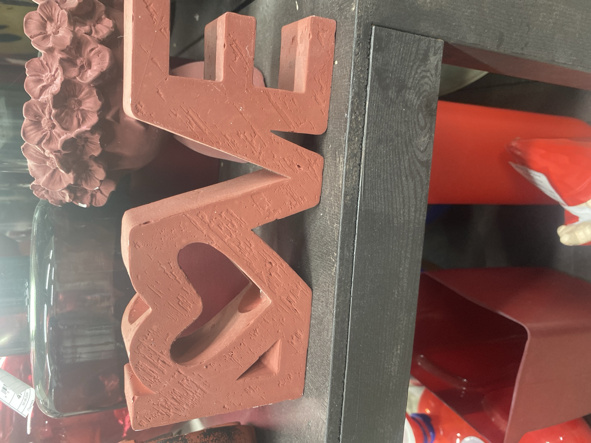 The image shows a close-up of the word "LOVE" spelled out in large, terracotta-colored, blocky letters. The 'O' is shaped like a heart. The letters are positioned vertically. To the left is a blurred floral arrangement in a glass vase. To the right, is a dark grey wooden shelf unit or display case. A few items can be seen on the shelves behind the word "LOVE", including a red canister, and red container. The photo appears to have been taken inside a store, possibly in Yekaterinburg, Russia, as the image was sourced from that location.