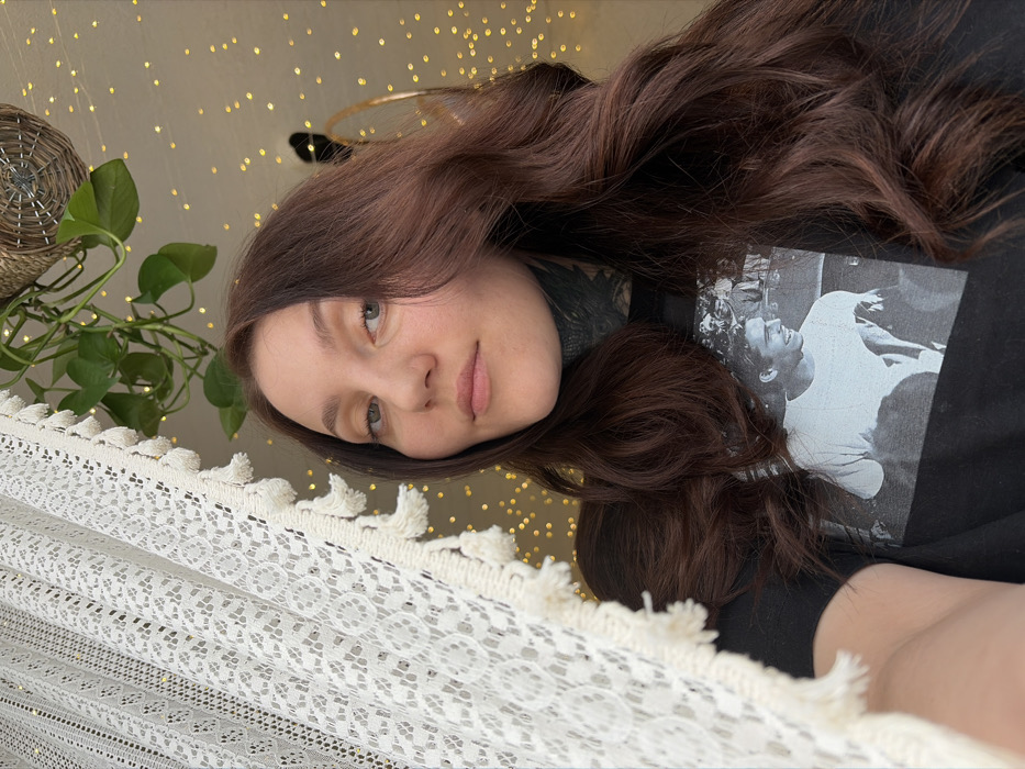 A young woman with long, wavy brown hair gazes at the camera with a slight smile. She wears a black t-shirt featuring a black and white image. A white, lacy fabric with tassels is draped in the foreground. Behind her, a green plant sits atop a woven basket, and string lights create a bokeh effect. A gold hoop is visible amidst the background clutter. The image has a slightly angled perspective.