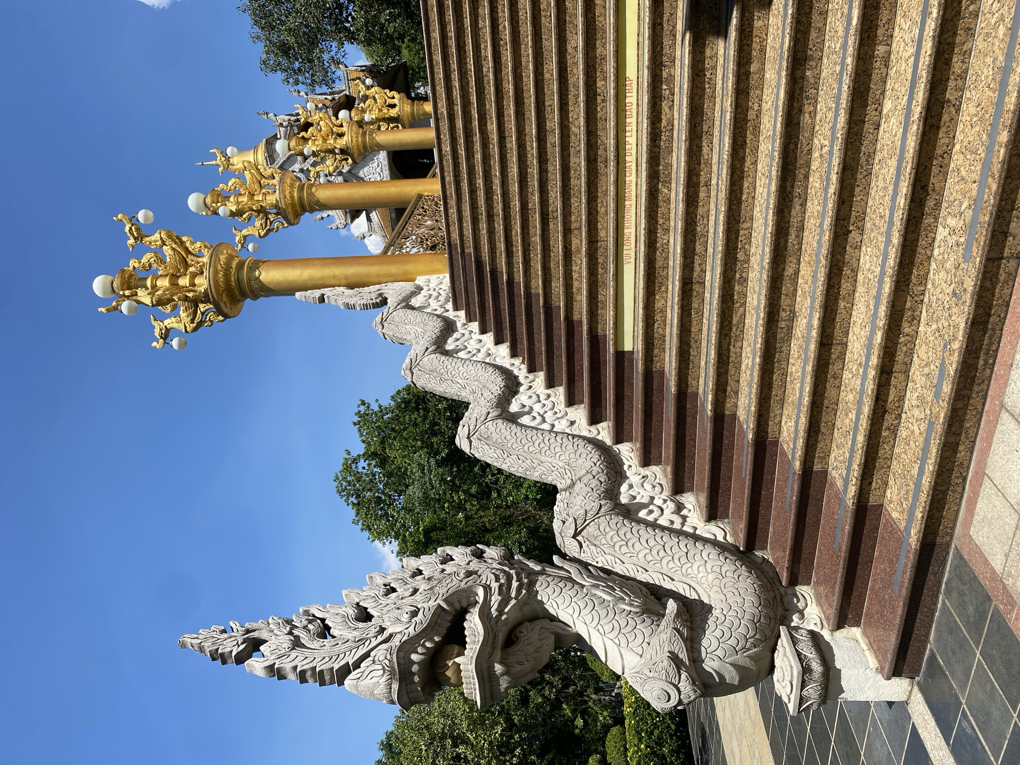 A view of a temple in Dĩ An, Vietnam presents a stone dragon-shaped statue that guards a staircase with granite stairs. Ornate gold lampposts with white globe lights appear adjacent to the stairs. Lush greenery and a clear blue sky form the backdrop.