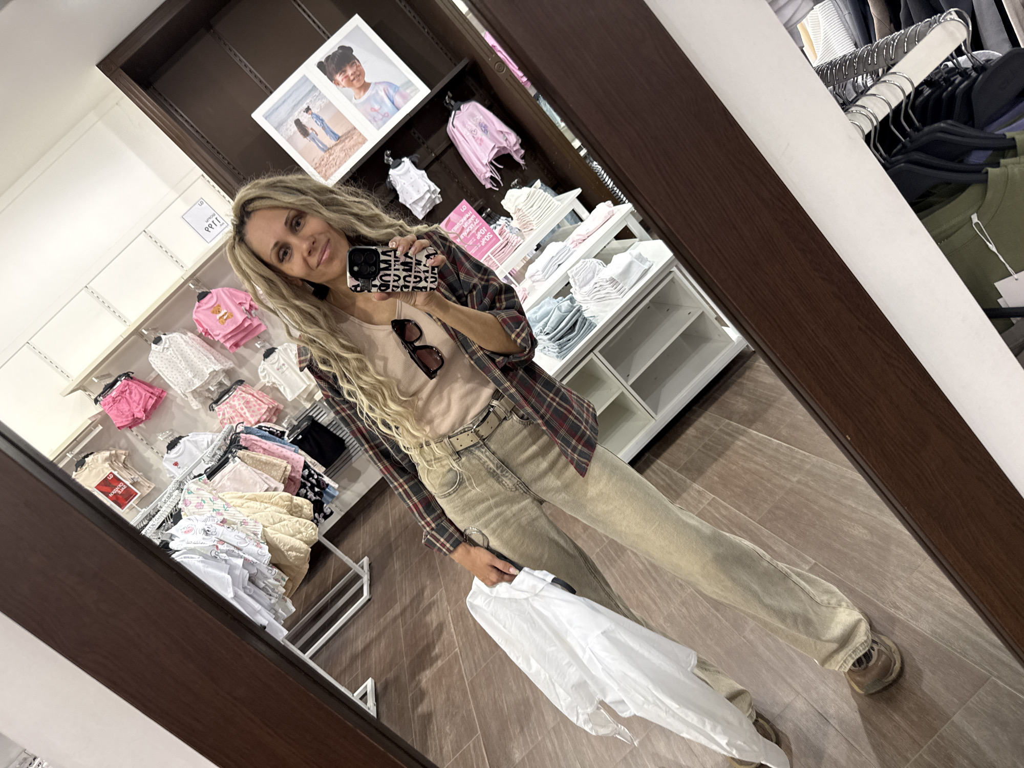 The image shows a woman taking a selfie in what appears to be a clothing store. The location is Unknown, Unknown. The woman has long, wavy blonde hair and is wearing a plaid shirt over a light-colored top and khaki pants. She is holding a white shirt in her hand and a phone in the other. Sunglasses are clipped to the front of her shirt. The store has racks of children's clothing on display and white shelves. The flooring is light-colored. A portrait is visible at the top of the background.