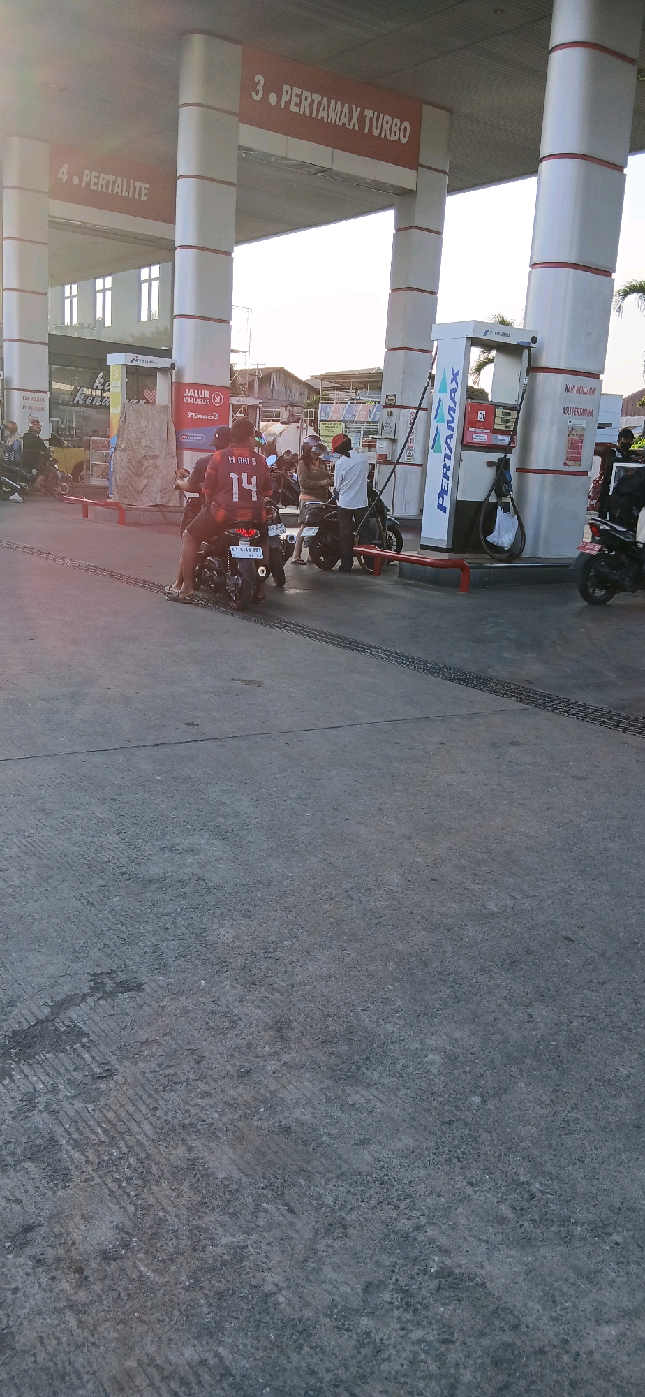 The image shows a view of a Pertamina gas station in Samarinda, Indonesia, likely during daylight hours. The gas station has multiple pumps and prominent signage indicating fuel types such as "4. PERTALITE" and "3. PERTAMAX TURBO." Several motorcycles are present, with a few people on or near them. One individual in the foreground is wearing a dark jersey with the number "14" and the text "MARI S" on the back. The ground is paved concrete, showing tire marks from vehicles. In the background, buildings and possibly other vehicles are visible. The overall scene depicts a typical operational gas station environment.