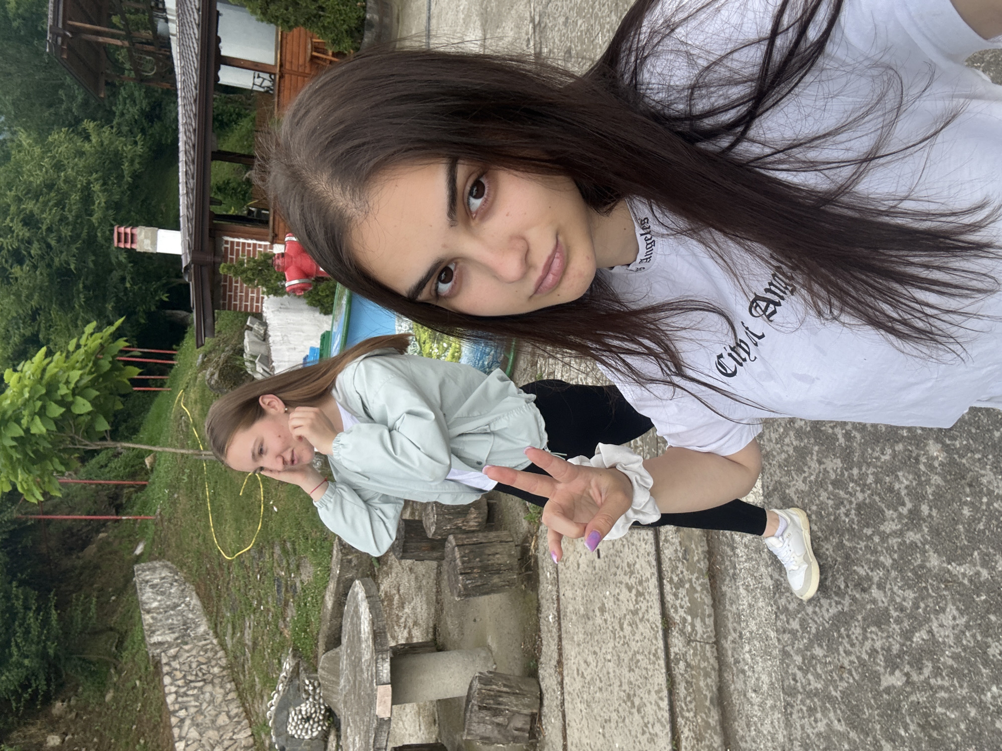 This media file captures two young women in an outdoor setting, likely a garden or yard, in Polena, Bulgaria. The image is oriented with the subjects and background rotated 90 degrees clockwise.
The primary subject, closer to the camera, has long, dark brown hair and is wearing a white t-shirt with the text "City Angeles" (or "City Angel") in black gothic font, black leggings, and white sneakers. She looks directly at the viewer with a slight smile and her right hand is raised, displaying a peace sign with light purple nail polish. A white scrunchie is visible on her wrist.
In the background, a second young woman with lighter brown hair is seen wearing a light green jacket over a white top. She has her hand near her face, possibly touching her ear, and her expression suggests a casual, perhaps contemplative, mood.
The scene is set on a concrete ground surface with a rustic outdoor seating area made of concrete and wooden stumps. Behind the second woman, a grassy slope with lush green foliage, trees, and bushes is visible, along with a yellow garden hose. In the far background, traditional-style buildings with brown roofs, a white chimney, and a brick wall section are discernible. A red object resembling a fire hydrant is also present near a building. The lighting indicates it is daytime, possibly with overcast skies, creating a soft, even illumination. The overall atmosphere appears relaxed and casual.
