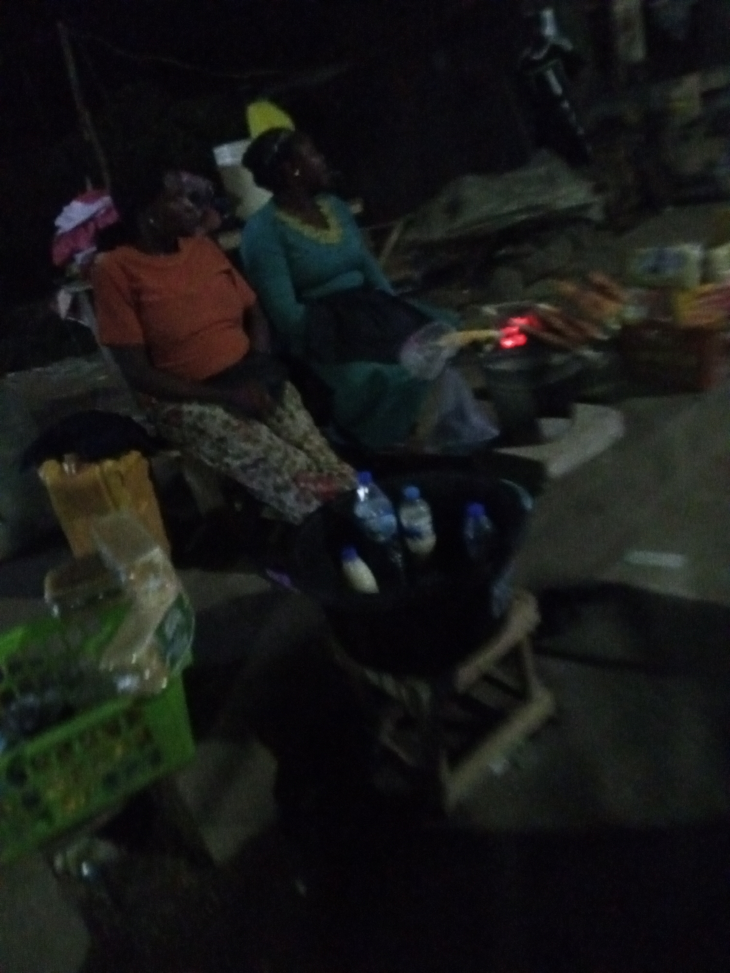 Two women are seated in an outdoor setting, potentially a street market, in what appears to be Bukuru, Nigeria, at night. One woman is wearing an orange top and patterned pants. The other is wearing a green dress with yellow trim. They are sitting near a table or stand with various items for sale, including bottled water, food items, and a green crate of goods. In the background, there is evidence of a small fire and other vendors or market stalls. A motorcycle is partially visible in the background as well. The scene is dimly lit.