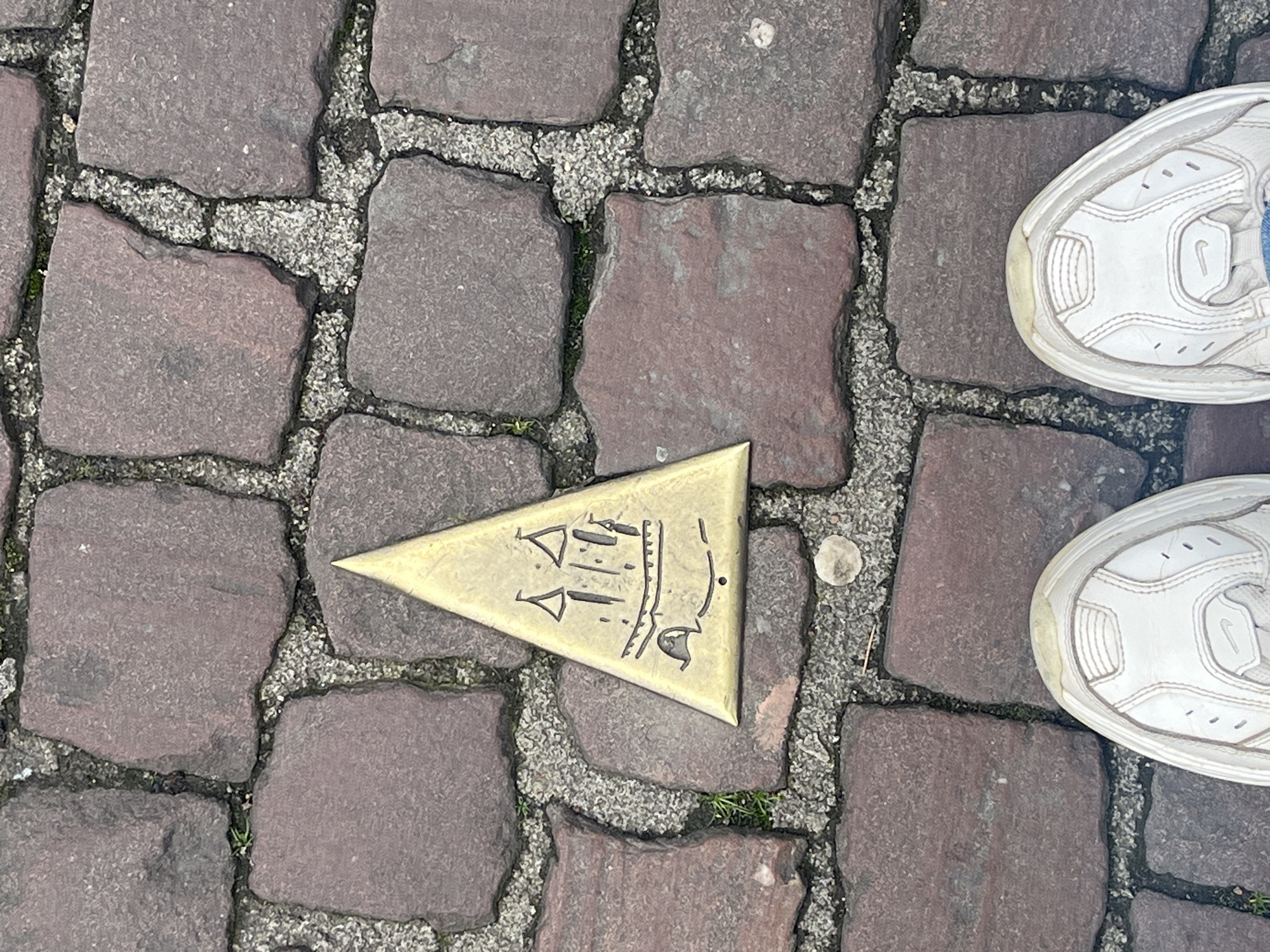 The image depicts a cobblestone street in Metz, France. In the center is a triangular brass plaque with an inscription pointing towards a specific location. A pair of white Nike sneakers are positioned on the right side of the image. The cobblestones are varying shades of red and gray. The brass plaque appears to be fixed into the pavement.