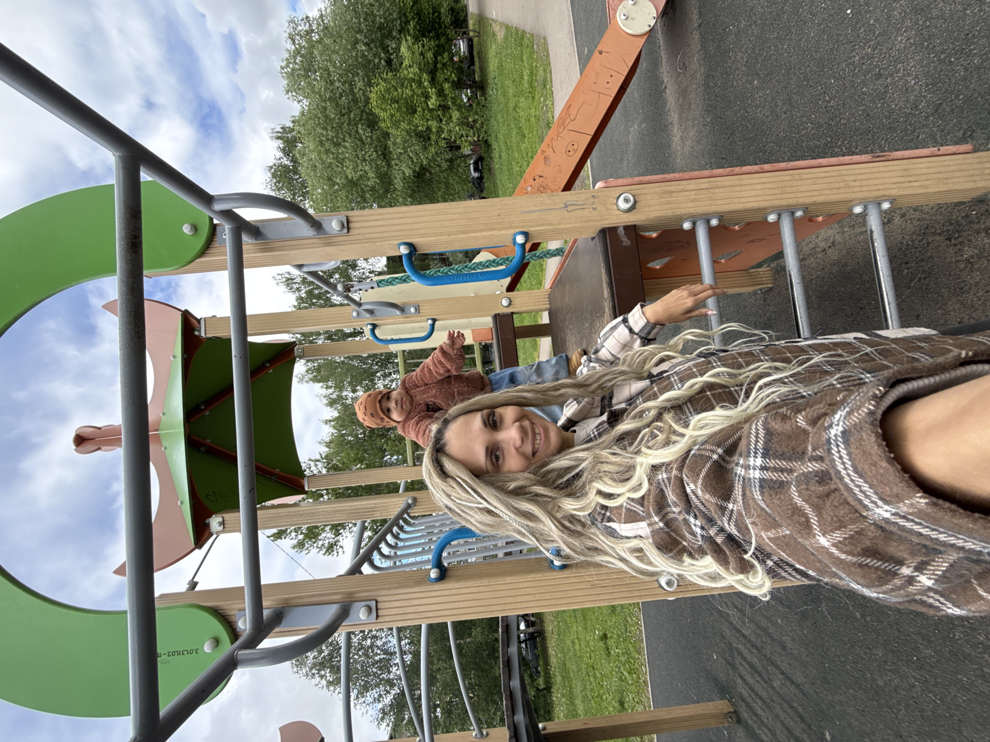 A woman with blonde hair and a brown plaid jacket smiles at the camera while sitting on playground equipment. In the background, a young child in a brown outfit and hat sits in a similar piece of playground equipment. The playground is located in Rusanovo, Russia. The scene includes elements of a playground, such as climbing structures, bars, and a slide. The background features greenery and a glimpse of the sky.
