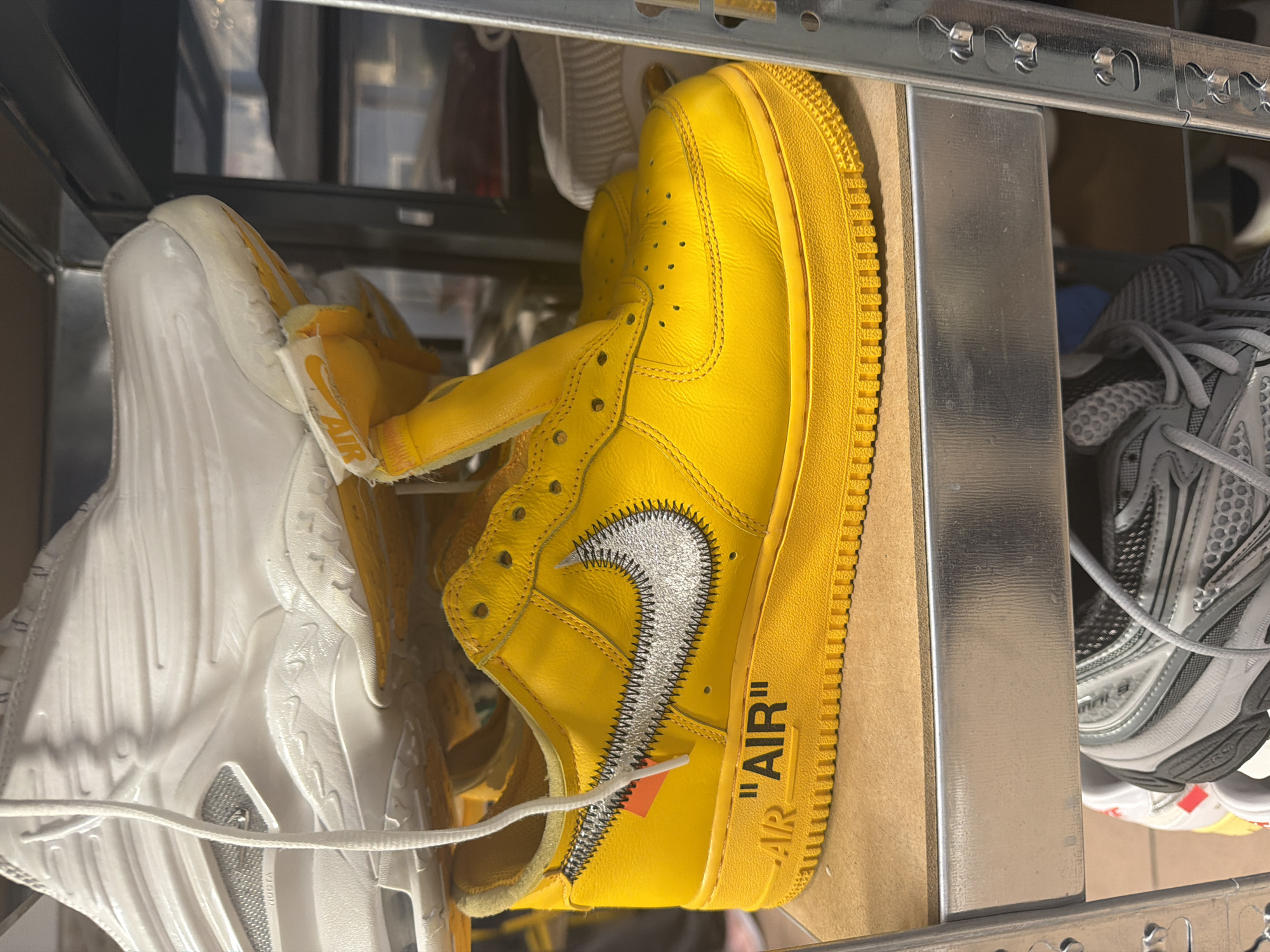 The image shows a close-up of a metal shelving unit filled with shoes. On the left is a pair of white Nike Air Foamposite sneakers. To the right, in the center of the frame, is a bright yellow Nike Air Force 1 sneaker with a silver Nike swoosh and the word "AIR" printed on the side. In the background, to the right, is a gray running shoe. The shoes are arranged on the shelves in a casual manner. Given the presence of Off-White branded Air Force 1, and in combination with Foamposites, the location is potentially in a retail store or a shoe collector's storage. Without further context, the exact location in Paris cannot be determined.