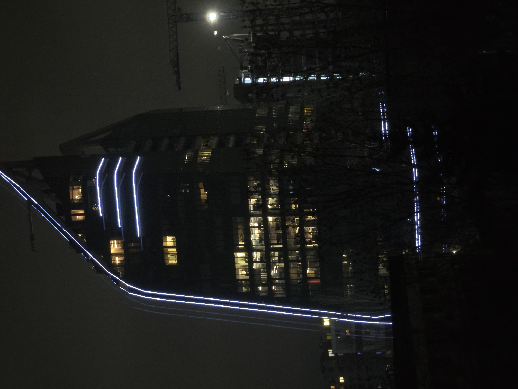 A nighttime view depicts a tall building with lights on in Sofia, Bulgaria. The building is illuminated with white vertical lights and yellow lights inside rooms. A crane is visible above the building. Trees and a dark area are in the right foreground.