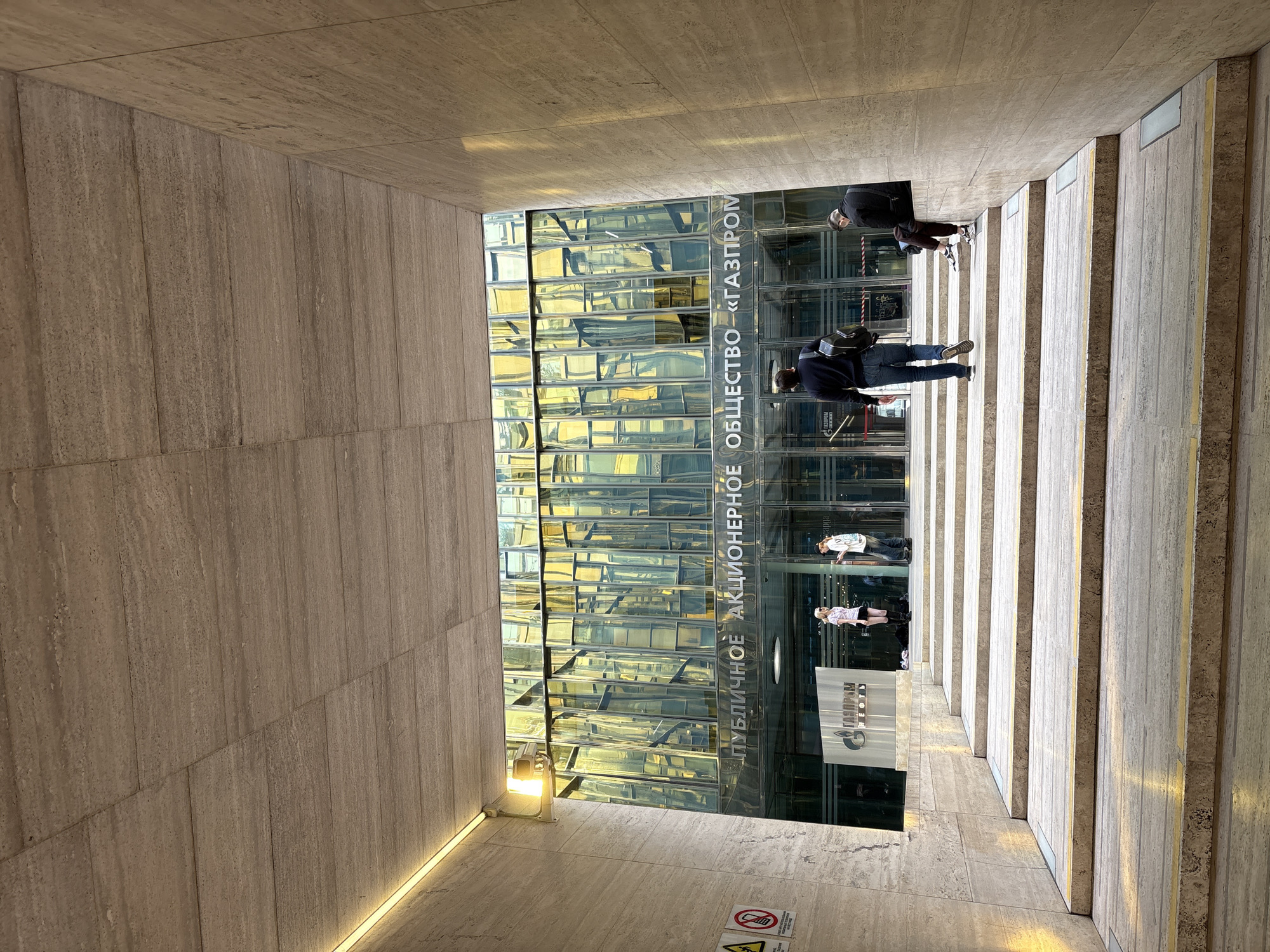 The image shows the entrance of the Gazprom headquarters in Saint Petersburg, Russia. The building is characterized by its glass facade reflecting sunlight. The entrance is framed by stone walls and steps, with several people walking in and out of the building. The building’s name “Газпром” (Gazprom) is visible on the facade. There are also some warning signs, and a narrow strip of LED lighting along the wall.