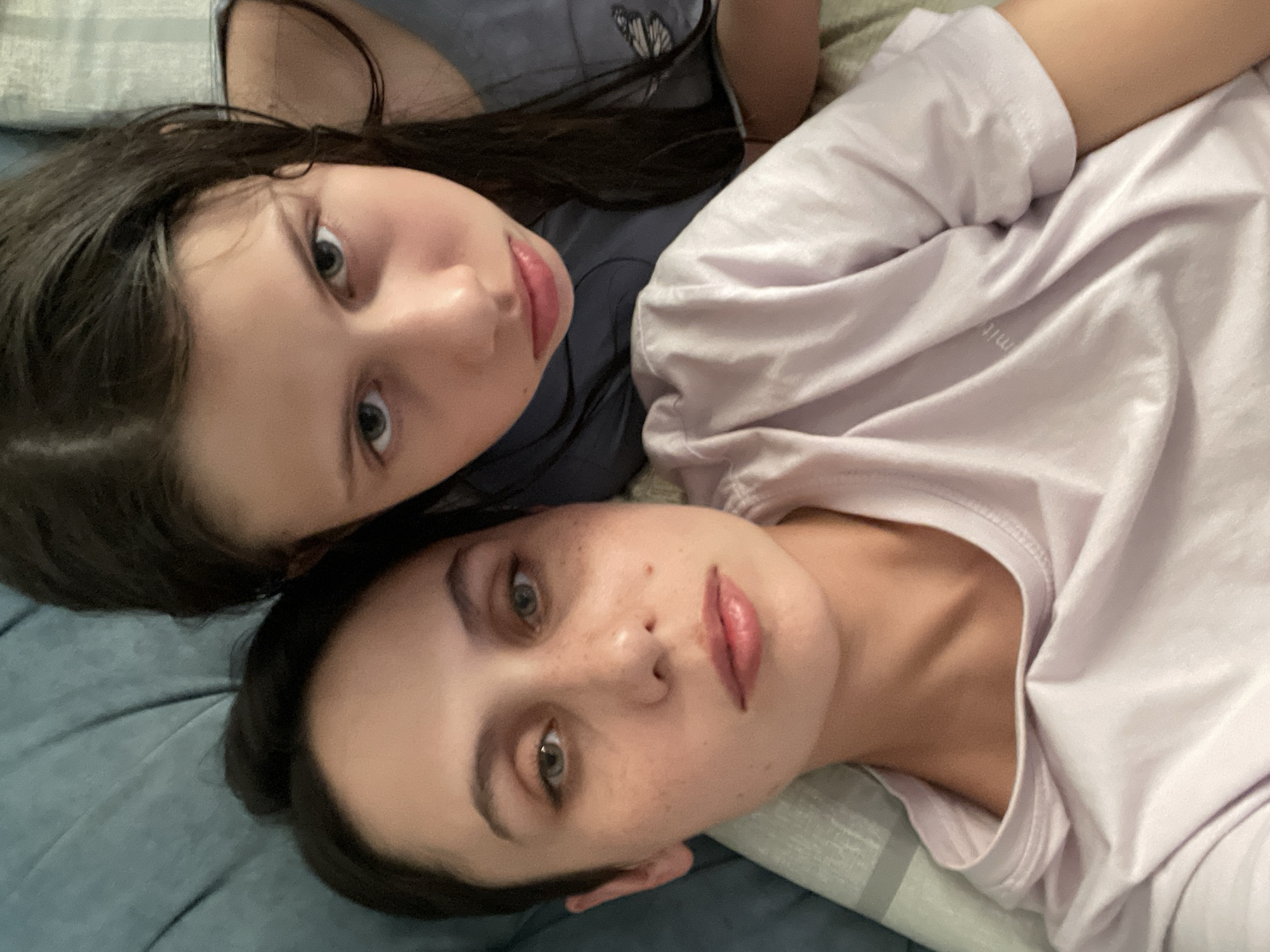 Two females are lying down, close together, facing the camera. One has dark hair and is wearing a blue top with a butterfly design. The other has dark hair, freckles, and is wearing a light purple t-shirt. They are on a blue and white patterned surface. The location is not identifiable as being in Izhevsk, Russia.