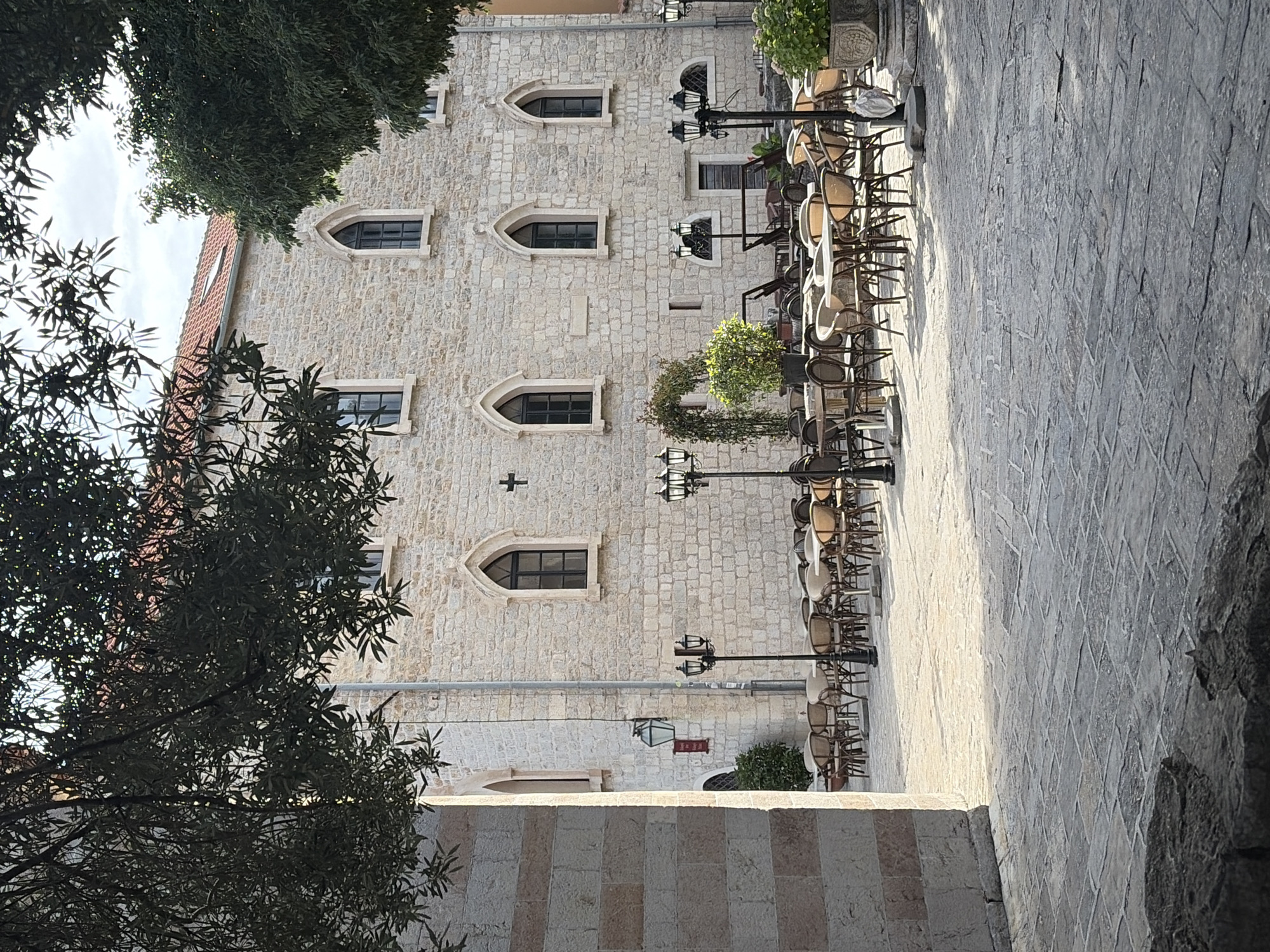 The image shows a narrow stone street in Budva, Montenegro, lined with a stone building featuring arched windows. Tables and chairs are arranged outside the building, suggesting a restaurant or cafe setting. Street lamps illuminate the area. Trees and foliage are visible in the foreground and on the building, partially obscuring the view. The ground is paved with stone tiles. The scene appears to be deserted at the moment the photo was taken.