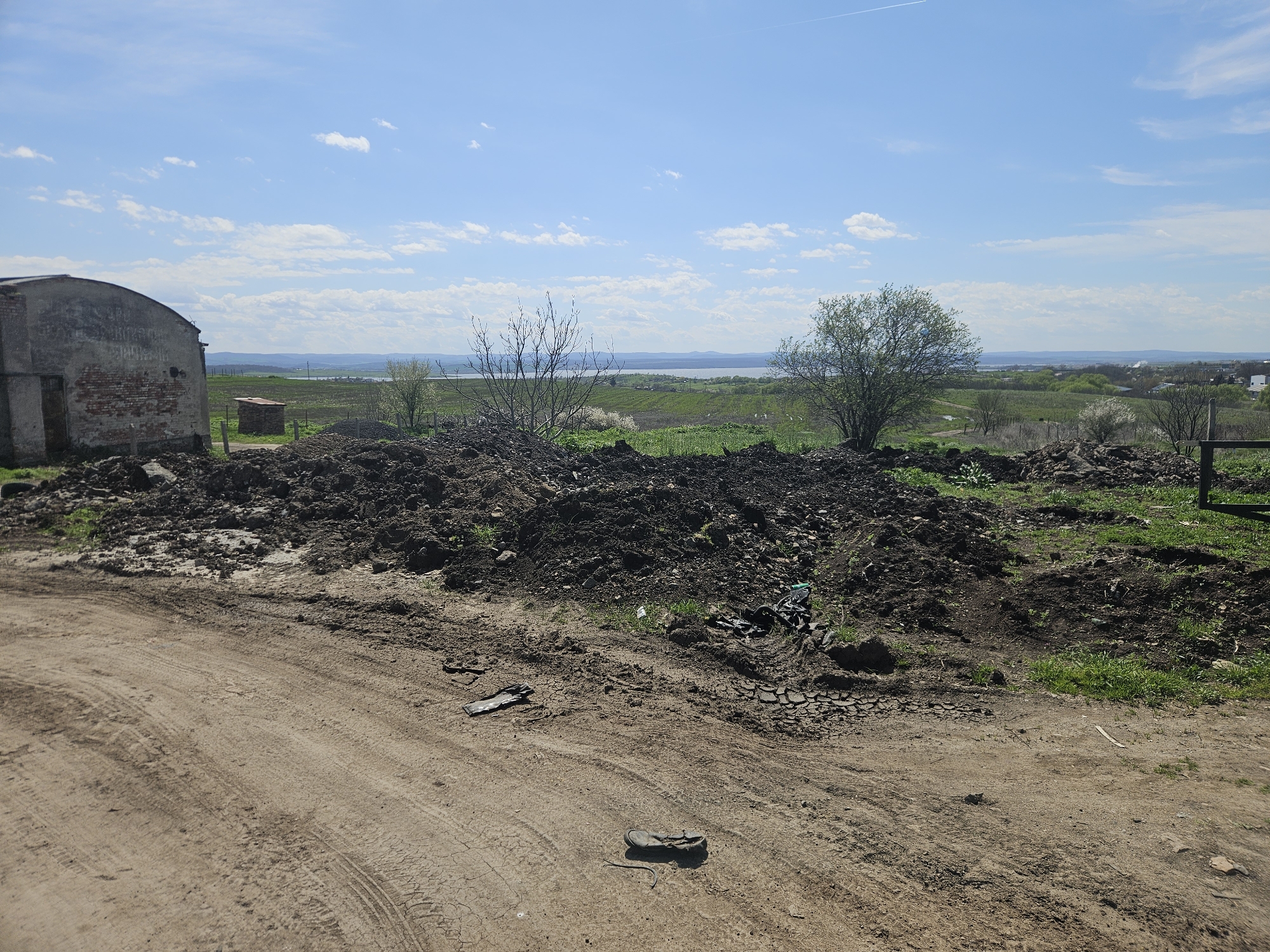 A landscape scene under a bright, partly cloudy blue sky shows a rural or semi-urban area in Burgas, Bulgaria. In the foreground, a dirt road marked with tire tracks leads toward a large pile of dark soil and debris. Scattered on the road and mixed into the soil are discarded items including what appears to be a shoe or slipper, pieces of plastic, and other unidentified trash. Behind the soil pile, vegetation, including grass, small trees, and bushes, extends towards the middle ground of the image. To the left of the scene is a dilapidated brick and concrete structure with a partially obscured entrance. In the distance, beyond the vegetation, is a visible body of water, possibly part of the Burgas Lakes, with hills and a settlement on the far shore. The overall scene suggests an abandoned or neglected area.