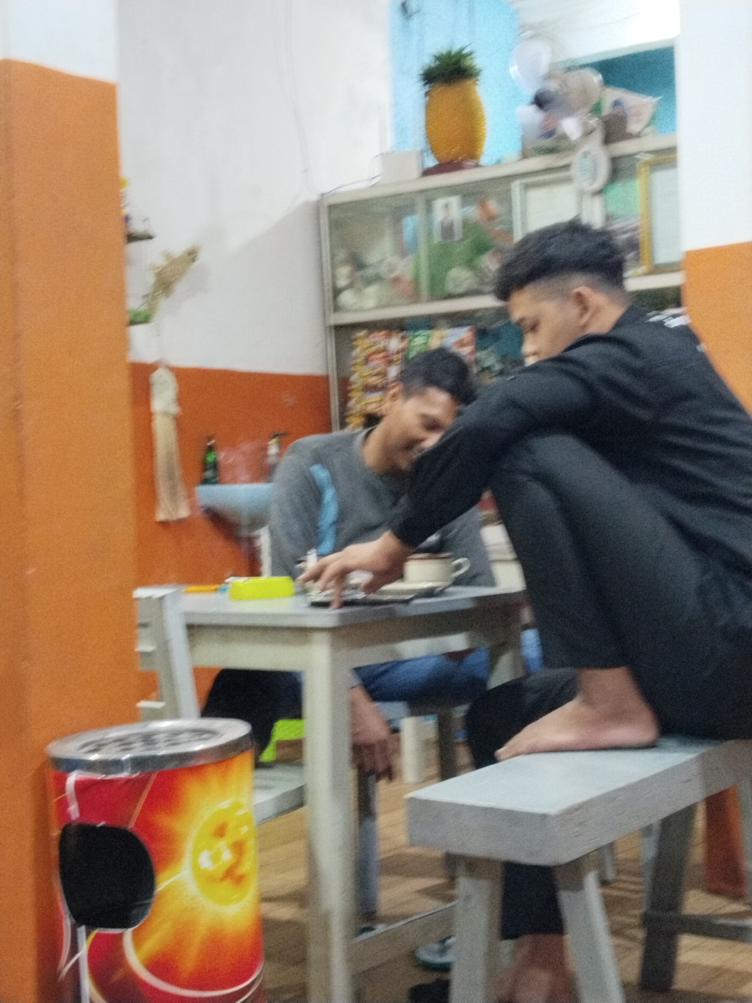 In the city of Prigi, Indonesia, a dimly lit indoor scene captures two young men at a table. The man on the right, wearing a black long-sleeved shirt and black pants, is seated with his legs crossed, leaning forward with his right foot resting on a light gray bench. His gaze is directed downwards. The man on the left, wearing a gray long-sleeved shirt, is also seated at the table, his head bowed as if engaged in a conversation or activity. A white mug and a small yellow object are visible on the table between them. In the background, a white cabinet displays various items, and a sink area is partially visible. To the left of the frame, a cylindrical trash can adorned with a sun motif stands prominently. The wall to the left is painted a vibrant orange, contrasting with the white walls elsewhere.