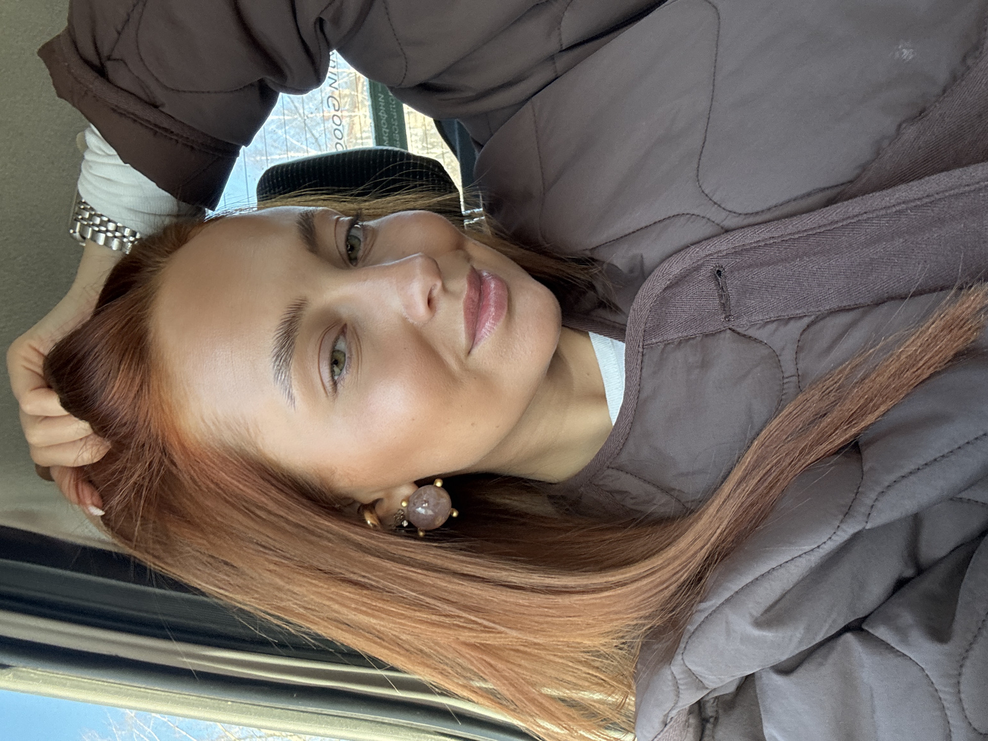 A close-up, tilted selfie captures a woman inside what appears to be a vehicle. She has long, straight, reddish-brown hair that falls over her shoulders. Her light-colored eyes (possibly green or hazel) look directly at the camera, and she has a soft, pleasant expression with a slight smile, accentuated by natural-looking makeup and rosy lips.
She is wearing a brown, quilted jacket over a white top. A silver-toned watch with a link bracelet is visible on her left wrist, and a large, round, pinkish-purple earring with gold prongs adorns her right ear. Her left hand is raised, gently touching her hair near her temple.
Through a window in the background, a clear blue sky and bare tree branches suggest a daytime setting, likely late autumn, winter, or early spring. Partially visible text on a sign or building reads "IN GOOD". The scene is set in Chaikovsky, Russia.