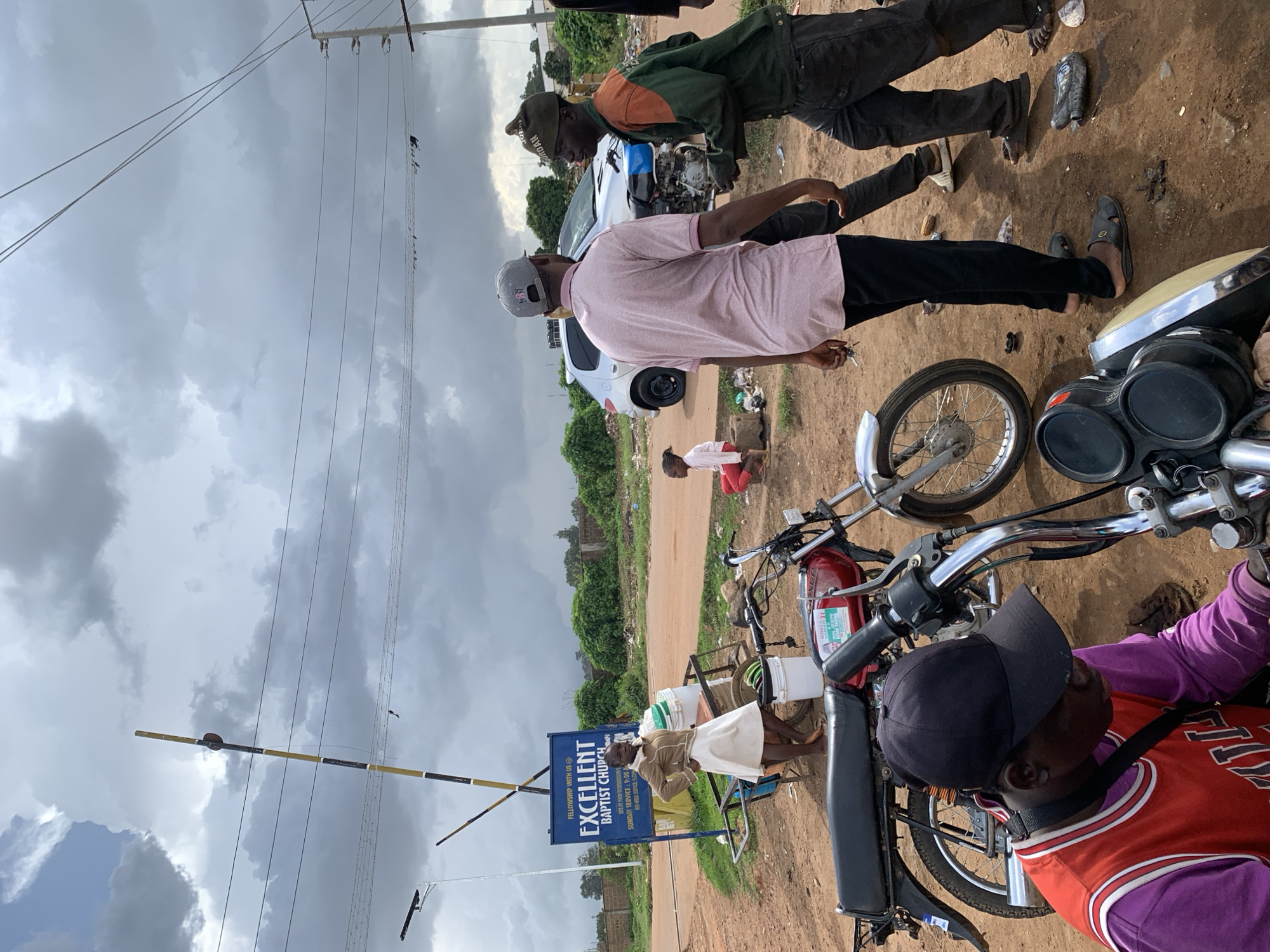 In Barakin Lerawi, Nigeria, a street scene unfolds under a cloudy sky. A young woman in a white skirt and brown jacket stands near a blue sign for "EXCELLENT BAPTIST CHURCH." Adjacent to her is a cart with buckets. In the foreground, two red motorcycles are parked on a dirt road. A man in a pink shirt and black pants, wearing a grey baseball cap, stands facing a man in a green and orange jacket and dark pants, wearing a green cap. The man in the pink shirt holds keys in his hand. A young girl in a pink shirt and red shorts sits to the left, while a child in a pink top and white shorts is visible in the middle ground. A silver car is parked behind the men. The environment is characterized by a dirt road, scattered debris, and lush green vegetation in the background. Power lines are visible overhead.