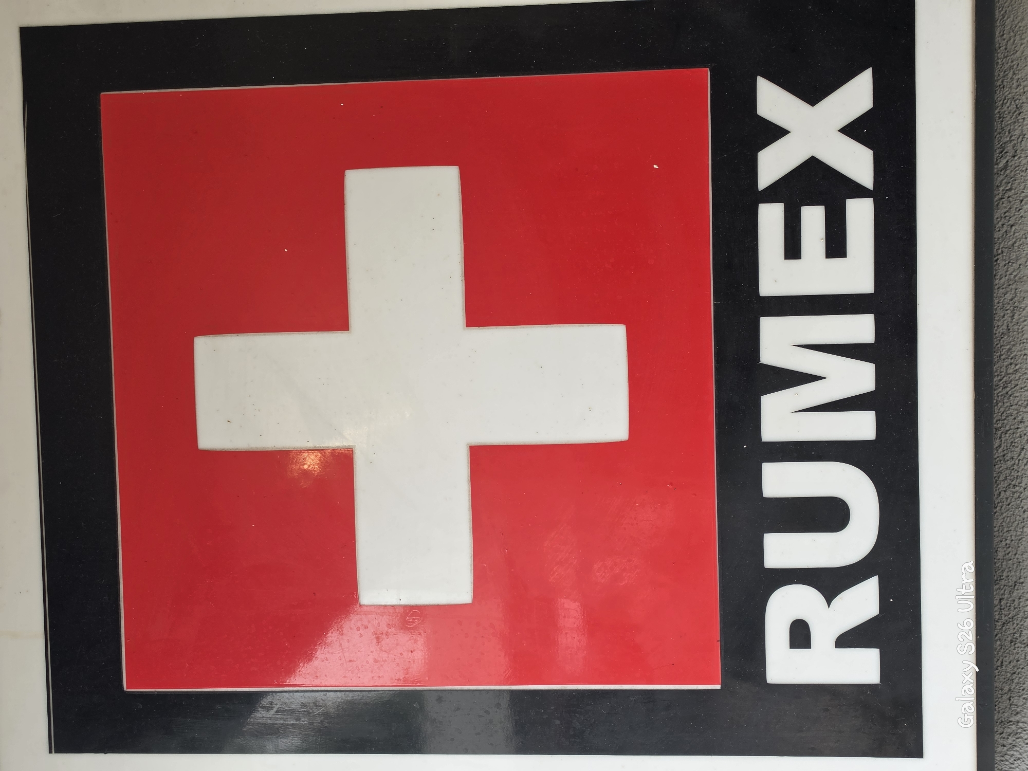 A close-up, slightly angled shot captures a sign with a red square in the upper portion, featuring a white cross, resembling the Swiss flag. Below the red square is a black rectangular area with the word "RUMEX" in white, bold, sans-serif capital letters. The sign has a white border. The background is out of focus, appearing to be a textured surface, possibly a wall or pavement. A subtle watermark "Galaxy S26 Ultra" is present in the lower left corner. The scene does not depict any people or discernible actions, and the specific location in Plovdiv, Bulgaria, is not evident from the visual information provided.
