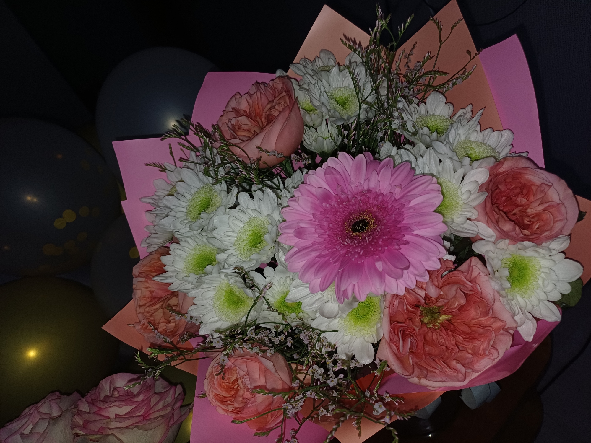 A close-up, dimly lit photograph showcases a bouquet of flowers. The bouquet is arranged with a variety of blooms, including a large pink gerbera daisy at the center, surrounded by white chrysanthemums with bright green centers. Several pink and coral-toned roses are interspersed throughout the arrangement, some in full bloom and others still tightly furled. The flowers are wrapped in pink and peach-colored paper, creating a soft, complementary backdrop. Delicate sprigs of tiny, light purple flowers and green foliage add texture and depth to the bouquet. In the background, out of focus, are several dark gray and gold balloons, suggesting a celebratory occasion. The lighting is low, with some areas of the image appearing dark, while the flowers and balloons are illuminated, creating a moody and intimate atmosphere. The overall impression is one of a festive, yet subdued, celebration. There is no visible text in the image.