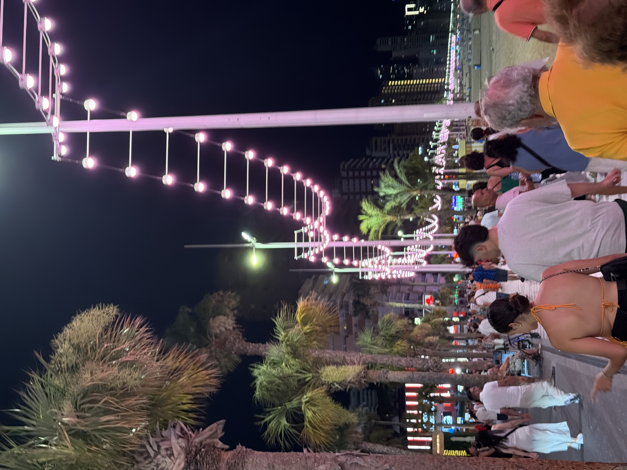 This is a nighttime, eye-level photograph of a pedestrian promenade in Benidorm, Spain. Palm trees line the walkway, their fronds illuminated by decorative string lights in shades of pink and white that are suspended from tall poles. In the background, multi-story buildings with illuminated windows are visible, indicating an urban setting. Numerous people are walking along the promenade, some in casual attire, while others are seated in a wheelchair. The atmosphere appears lively and social, suggesting a popular tourist area.