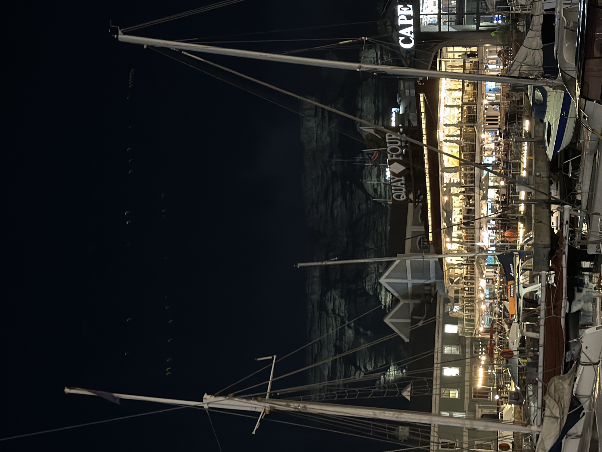 The image shows a nighttime scene at the V&A Waterfront in Cape Town, South Africa. In the foreground are masts and rigging of several sailboats docked in the harbor. The masts reach into a dark night sky. The water reflects the lights from buildings. A building with the signage "QUAY FOUR" and "CAPE" is illuminated, showing several levels of a restaurant bustling with people. The background shows a dark, rocky cliff face, likely part of Table Mountain. The overall scene captures a vibrant nightlife atmosphere with the combination of boats and restaurants.