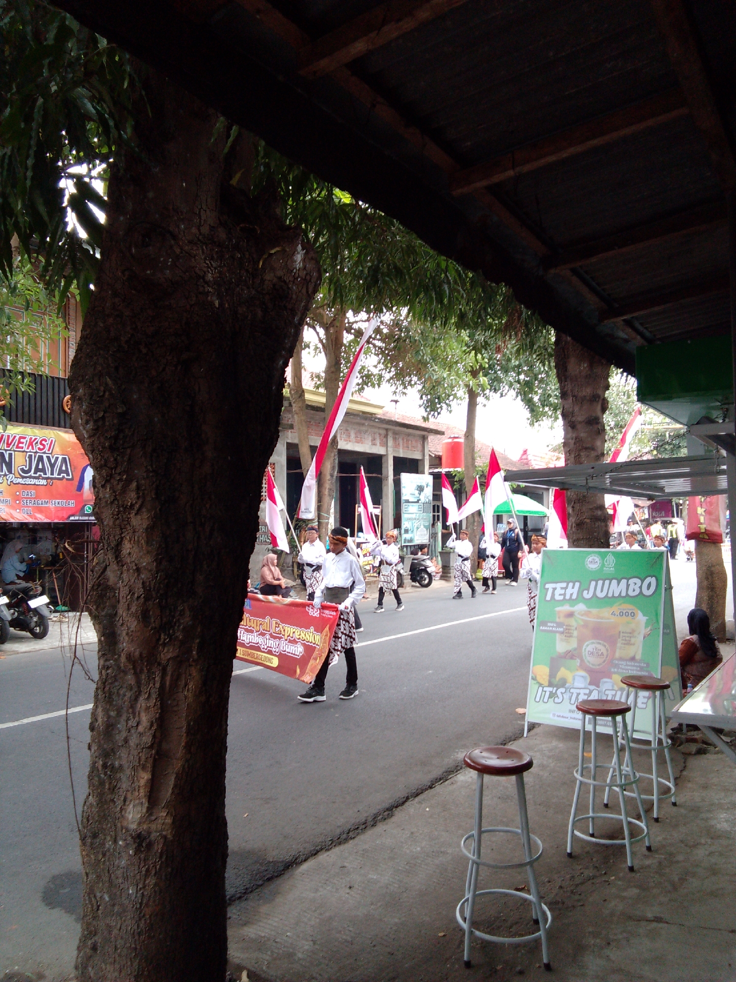 In Trenggalek, Indonesia, a parade or procession is underway on a street. A large tree trunk dominates the left foreground. Several people in traditional clothing, including men in white shirts and patterned sarongs, are walking in the procession, carrying a red banner that reads "Cultural Expression". Indonesian flags are also visible, held by participants and planted along the street. In the background, buildings and shops line the road, one of which has a banner with "N JAYA" and other text. A green sign advertising "TEH JUMBO" with a price of 4.000 is prominently displayed on the right side of the image. Three bar stools are placed on the sidewalk in front of the sign. A woman wearing a hijab is seated near the sign. The scene appears to be a daytime event with moderate activity.