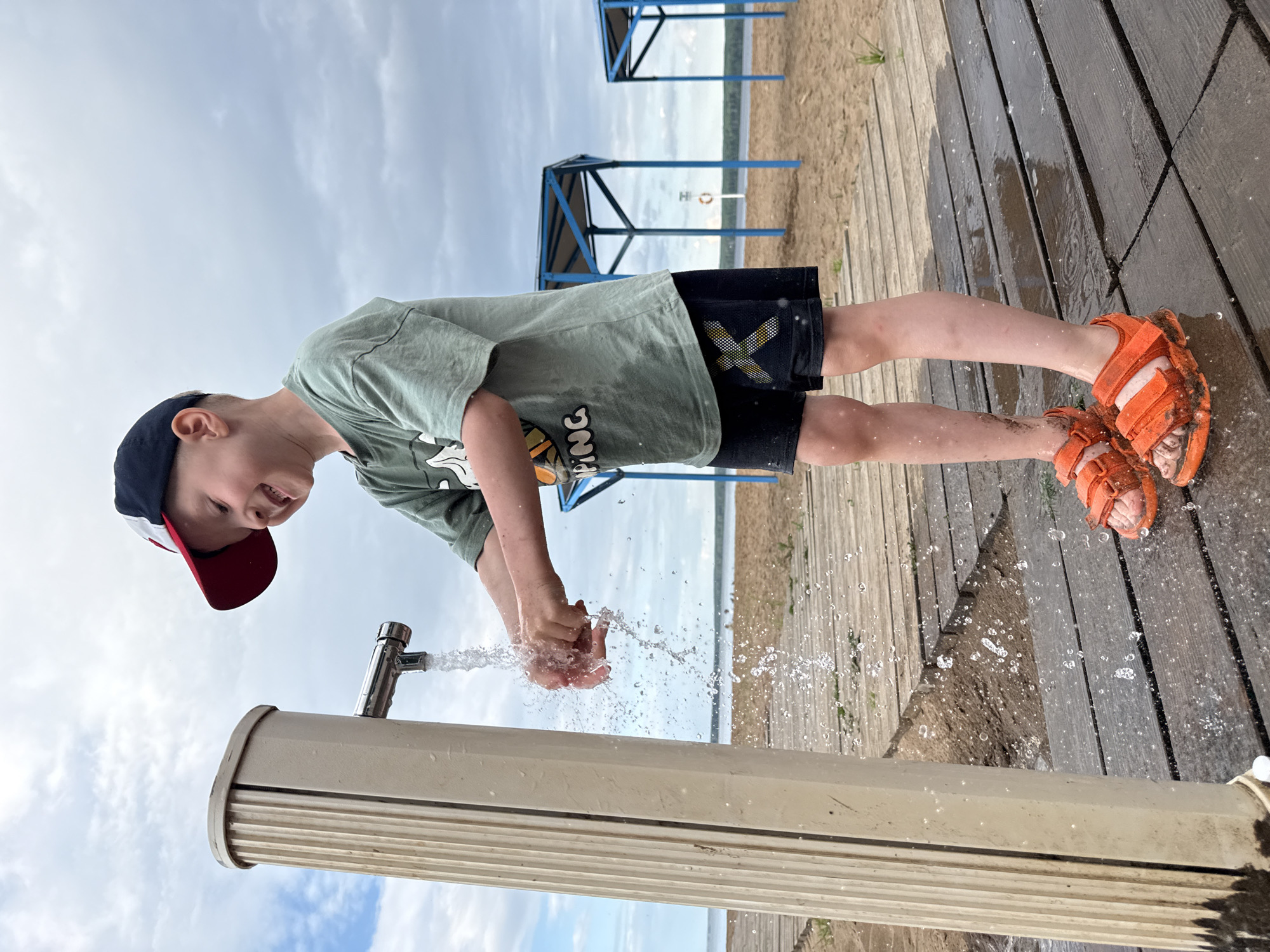 A young boy wearing a green t-shirt, black shorts with a yellow 'X' logo, a baseball cap, and orange sandals is washing his hands under a water spigot at a beach in Chaikovsky, Russia. He is standing on a wet wooden boardwalk. In the background, there is a sandy beach, the Kama River, and two blue structures that appear to be shelters. The sky is overcast with clouds. The scene appears to be a public area near a body of water.