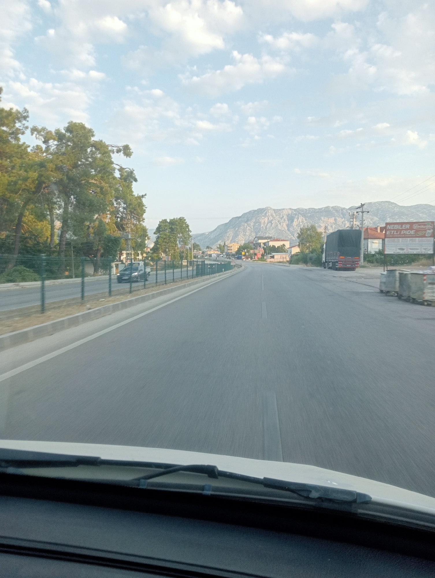 The image depicts a view from the windshield of a vehicle driving on a paved road in Döşemealtı, Turkey. The road stretches into the distance, with a clear lane marking dividing the direction of travel. On the left side of the road, a green metal fence runs alongside, separating it from a strip of greenery and mature trees. Beyond the trees, residential buildings are visible. On the right side of the road, a large truck with a dark green tarp covering its cargo is parked. Adjacent to the truck, there is a billboard with Turkish text that reads "NEBİLER ETLİ PİDE". Further down the road on the right, more buildings and what appear to be large industrial bins are present. In the background, a mountain range rises, its slopes etched with geological detail. The sky is a pale blue, dotted with scattered white clouds. The scene appears to be captured during daylight, likely in the late afternoon or early morning, given the soft lighting. No people are directly visible in the image, although the presence of vehicles and buildings suggests human activity in the area.