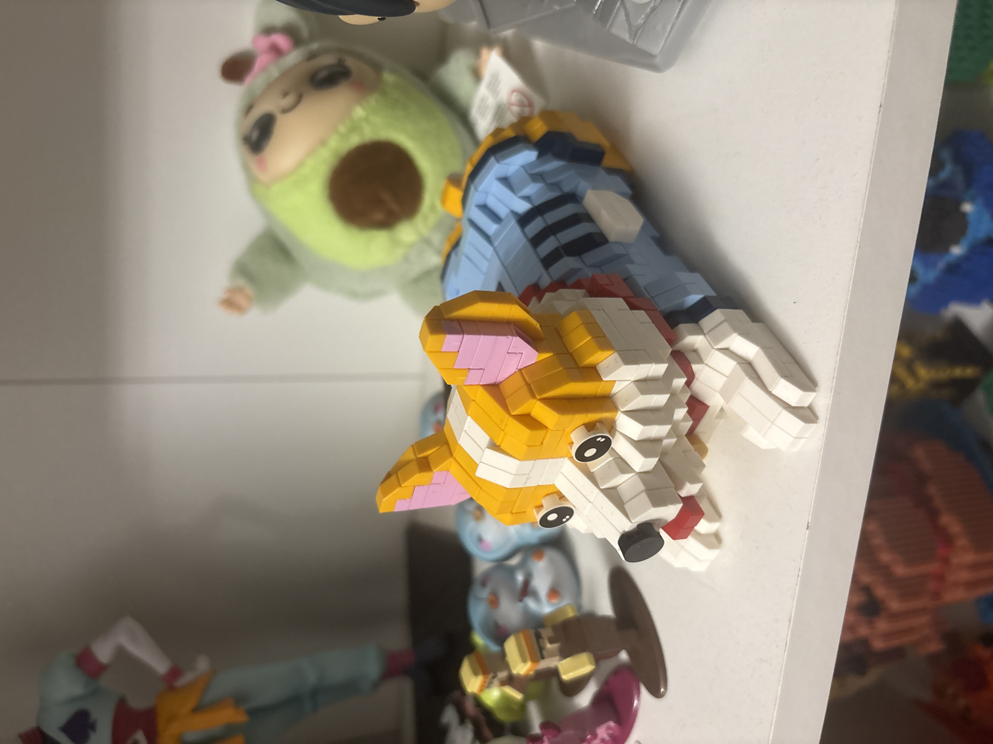 The image shows a shelf containing various toys and figurines. In the upper left, there's a green avocado-shaped plush toy with a face. Beside it is a Lego-like figurine of a corgi dog wearing a blue shirt.
Below, partially visible, are other toys. There is no information in the photo about the location. There is no evidence to suggest this picture was taken in Plovdiv, Bulgaria.