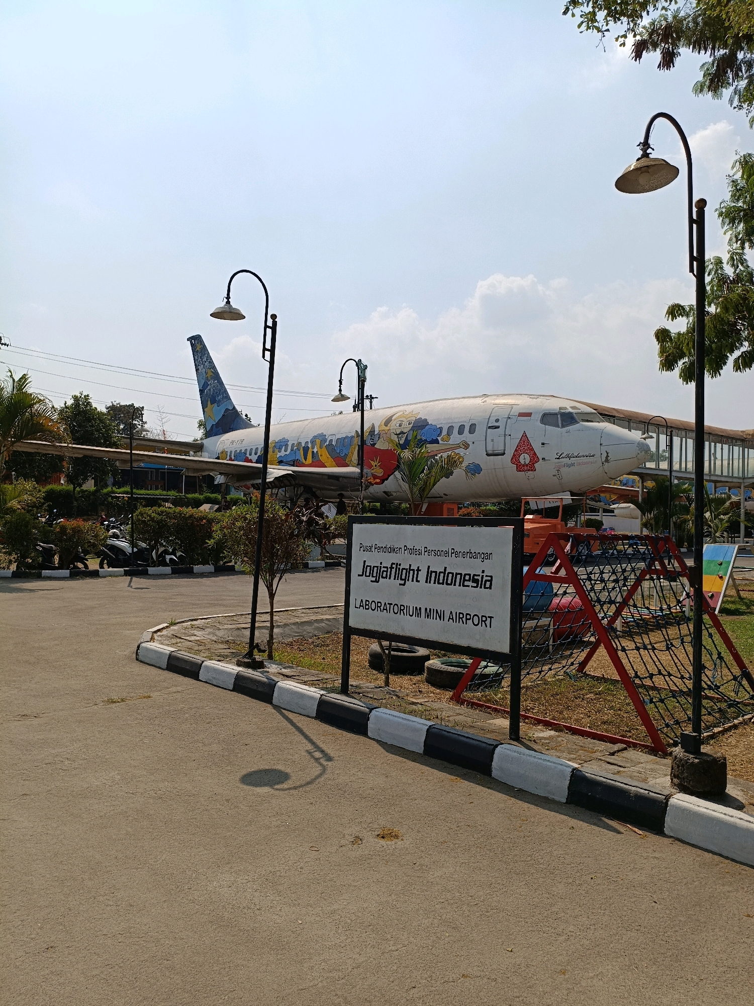 The image shows an outdoor scene in Purwomartani, Indonesia, featuring a decommissioned airplane used as an exhibit or attraction. The airplane, a narrow-body jet with a white fuselage adorned with colorful cartoon characters, is parked on a tarmac. In the foreground, a paved pathway curves to the left, bordered by a curb painted with alternating black and white stripes. Several street lamps with curved arms are visible throughout the scene. A sign in front of the airplane reads "Pusat Pendidikan Profesi Personel Penerbangan Jogjaflight Indonesia LABORATORIUM MINI AIRPORT". To the right of the sign is a children's climbing structure made of ropes and red metal bars. Behind the airplane, a walkway or bridge structure is visible. Parked motorcycles are present in the background, near a line of bushes and trees. The sky is partly cloudy and the sun is shining, casting shadows on the ground. The overall impression is of a recreational or educational facility centered around aviation.