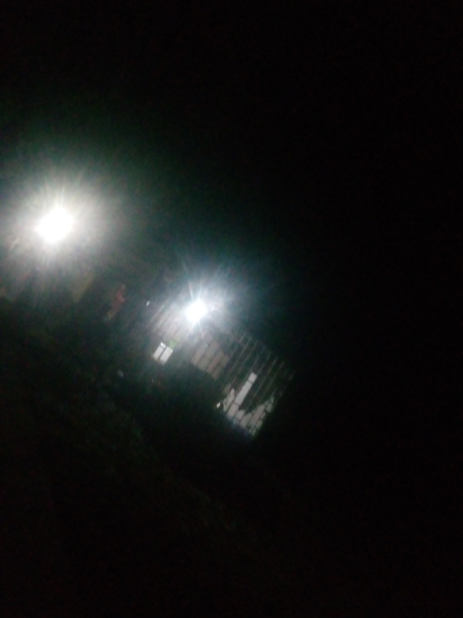 The image depicts an outdoor scene at night, characterized by low light conditions and significant glare from artificial light sources. Two prominent bright lights, likely streetlights or floodlights, illuminate portions of the scene. In the foreground, a dark, textured surface, possibly a wall or ground, is visible. Beyond this, there appears to be a structure with some fencing or grates, and possibly windows. The overall impression is that of a dimly lit urban or semi-urban environment, with artificial lighting struggling to overcome the darkness. There is no clear indication of the specific location within Jalingo, Nigeria, beyond the context provided in the prompt. No people are clearly discernible in the image.