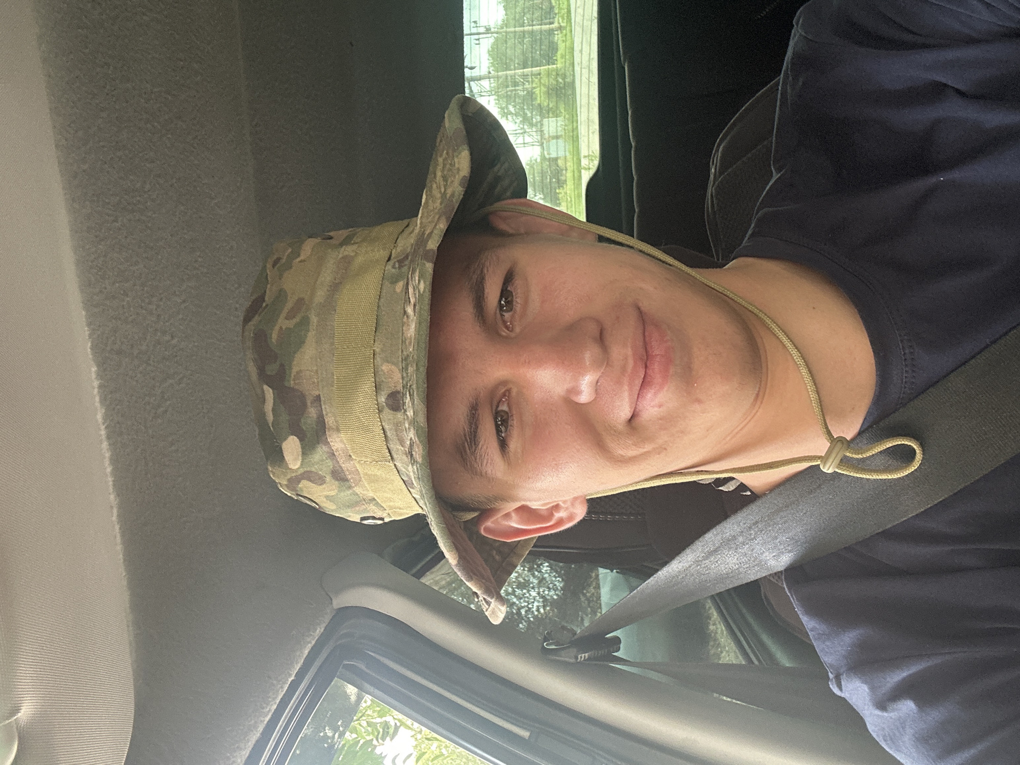 A young adult male is pictured from the chest up, wearing a camouflage bucket hat and a dark blue t-shirt. He is inside a vehicle, with a seatbelt visible across his chest and shoulder. The man is looking directly at the camera with a slight smile. The background shows the interior of the car, including the car's ceiling and a glimpse of the outdoors through the windows. The man's clothing and hat suggest an outdoor or potentially military-related context. The outdoor scenery visible through the windows shows greenery and what appear to be structures, possibly in a suburban or rural area. There is no specific visual information in the image that allows for the identification of the location as Oral, Kazakhstan.