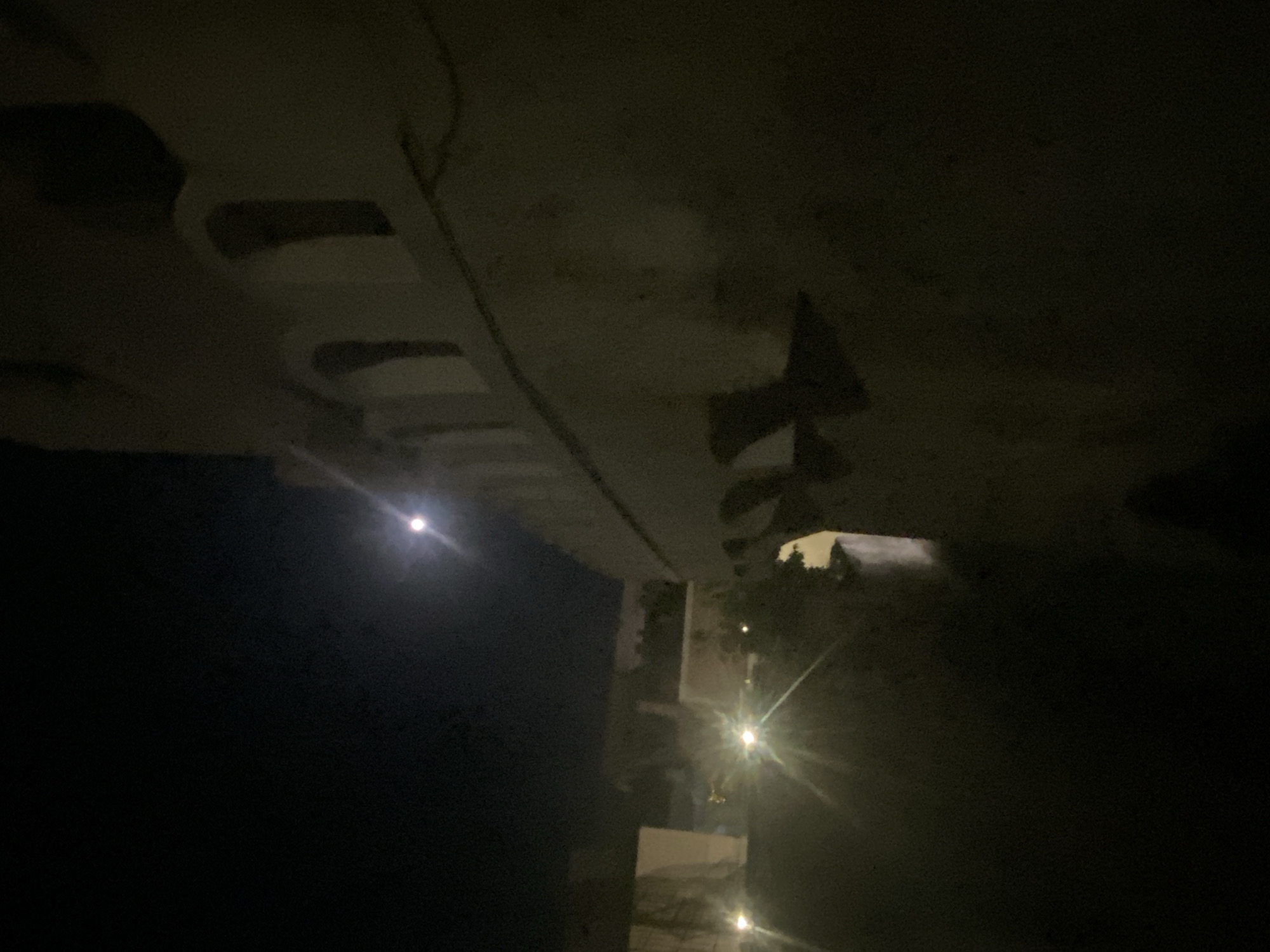 This photograph captures an outdoor nighttime scene, likely in the municipal unit of Thira, Greece. The dominant feature is the illuminated facade of a building, possibly a traditional Cycladic structure, characterized by arched openings and a textured, light-colored surface. The building appears to be a fortification or an older, substantial construction.
The scene is illuminated by artificial lights, evidenced by several bright light sources that create starburst effects and cast long, dramatic shadows. One prominent light source is on the left side of the frame, projecting rays outwards. Another light is visible further back, also illuminating parts of the building and surrounding vegetation.
The sky above is dark, suggesting late evening or night. A bright, round celestial body, most likely the moon, is visible in the upper portion of the image, slightly to the left of the center. Its luminescence is softened by the atmospheric conditions or distance.
The foreground is largely dark and indistinct, with some hints of paved ground or a pathway. The overall impression is one of a historical or atmospheric location under the cover of night, with man-made lights and natural moonlight contributing to the ambiance. There are no people or specific actions depicted in the photograph. The focus is on the architecture and the play of light and shadow in the nocturnal environment.