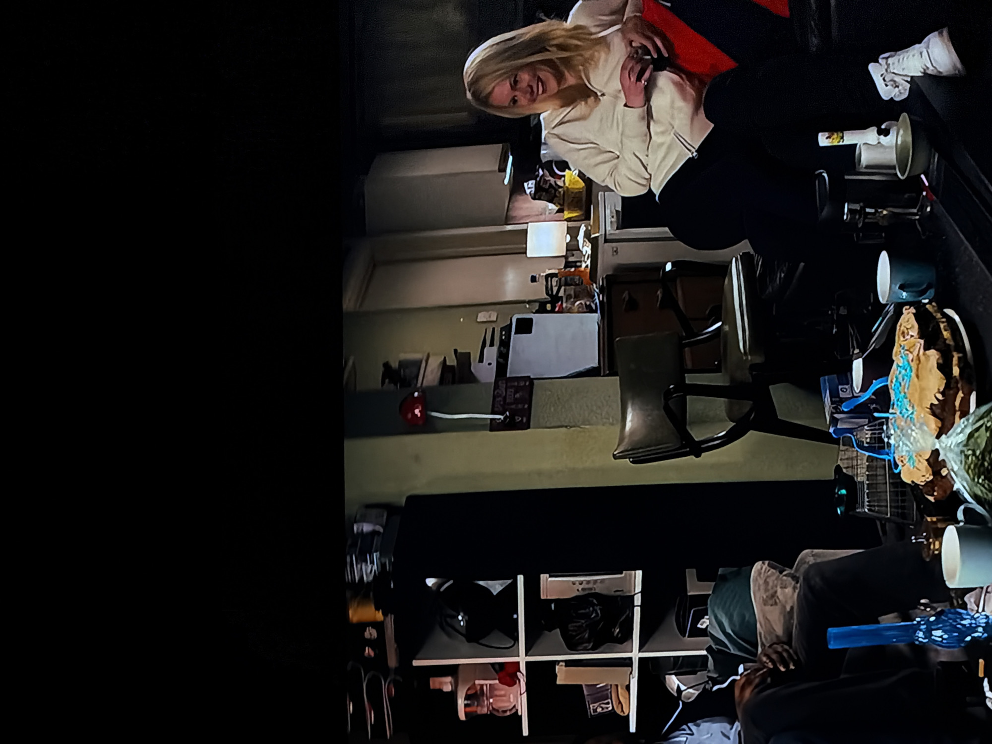 This image, located in Sofiivska Borshchahivka, Ukraine, depicts an indoor scene, likely a casual living space or room. The photograph is taken from a slightly high angle and is rotated 90 degrees clockwise.
The primary subject is a blonde woman in the upper central part of the frame, smiling and looking towards the viewer. She is wearing a cream-colored hooded sweatshirt and dark trousers, and appears to be sitting or leaning against something red, possibly a couch or chair. Her left hand is visible, holding a small object, possibly a phone.
To the right of the woman, various items are placed on what appears to be a dark table or surface, including a white boot or high-top sneaker, a white plunger, and a light-colored mug. Further down and to the right, a teal mug and a decorative item, possibly a cake or craft with blue and brown elements, are visible.
In the lower right corner, another person is partially visible, wearing a dark hoodie and appearing to be lying down or reclining.
The background reveals typical room furnishings. A shelf with several cubbies holds various items such as bags, shoes, and small appliances. A dark chair with metal legs is centrally located. In the middle-ground, a white lamp and various containers are visible on a surface. A red, wall-mounted device with a wire extends from the left side of the background.
The overall lighting suggests an indoor setting, possibly during the evening or night, creating some shadows but generally well-lit. The woman's smile indicates a positive and relaxed atmosphere. No discernible text is visible on any objects.