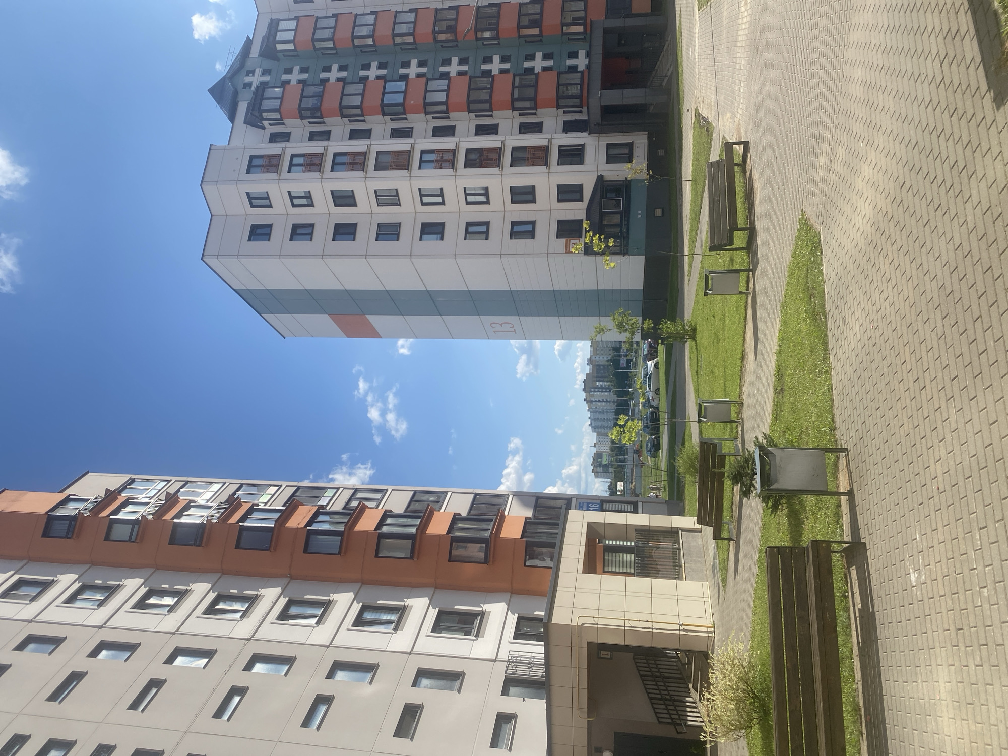 This is a photograph taken in Minsk, Belarus, featuring a residential courtyard with modern apartment buildings. In the foreground, a paved walkway made of interlocking bricks leads through a manicured green space with grass and a few young trees. Two dark wooden park benches are situated on the grass, each accompanied by a metal trash receptacle.
The main focus of the image is on the two tall, multi-story apartment buildings that dominate the skyline. The building on the left is predominantly cream-colored with vertical orange accents and numerous windows. It has a lower, beige-colored entrance structure with a staircase.
The building on the right is also modern, with a white facade and colorful vertical bands of teal and orange. It features many windows and balconies with dark railings. The number "13" is visible in a faded red color on the teal section of this building. Further in the background, more apartment buildings and some trees are visible under a bright blue sky with scattered white clouds. A street with parked cars can be seen between the courtyard and the distant buildings. The overall scene depicts a well-maintained urban residential area on a sunny day.