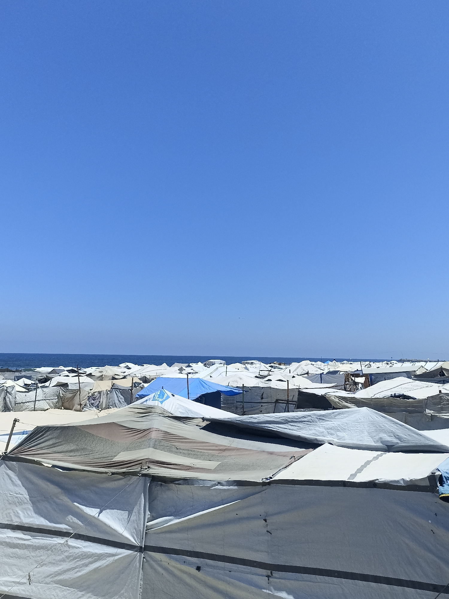 The image captures a sprawling refugee camp under a clear blue sky, likely during the daytime given the bright sunlight casting sharp shadows. The scene is dominated by rows upon rows of temporary shelters, predominantly white and gray canvas tents, constructed with poles and ropes. Some tents are pitched close together, creating a dense network of temporary housing, while others are more spread out. One prominent blue tent stands out amidst the neutral tones of the other shelters.
In the background, a thin line of the ocean is visible, suggesting the camp is situated near the coast. The water appears calm, reflecting the bright sky. The overall impression is one of a vast, makeshift community, likely established to house displaced people. The sheer number of tents and the simple construction materials evoke a sense of necessity and perhaps a precarious living situation. There are no visible people in the frame, but their presence is implied by the existence of the shelters. No text is discernible in the image. The setting appears to be a beach or sandy area, as indicated by the light-colored ground visible between the tents. The weather is fair and sunny, contributing to the starkness of the scene.