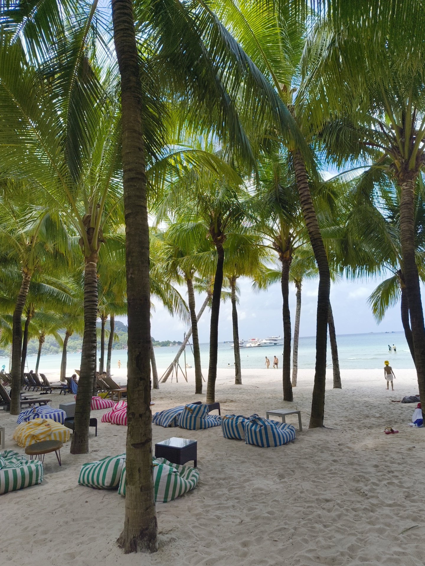 The image shows a beach in Phu Quoc, Vietnam, with numerous palm trees lining the sandy shore. In the foreground, several striped beanbag chairs and small tables are arranged on the sand, suggesting a lounge area. Beyond this, beach chairs are visible. Further out, people are seen walking along the shoreline and wading in the turquoise water. A few boats, including a larger white vessel, are anchored in the calm sea. The background features lush green hills and a clear blue sky with some clouds. The scene depicts a relaxed tropical beach environment.