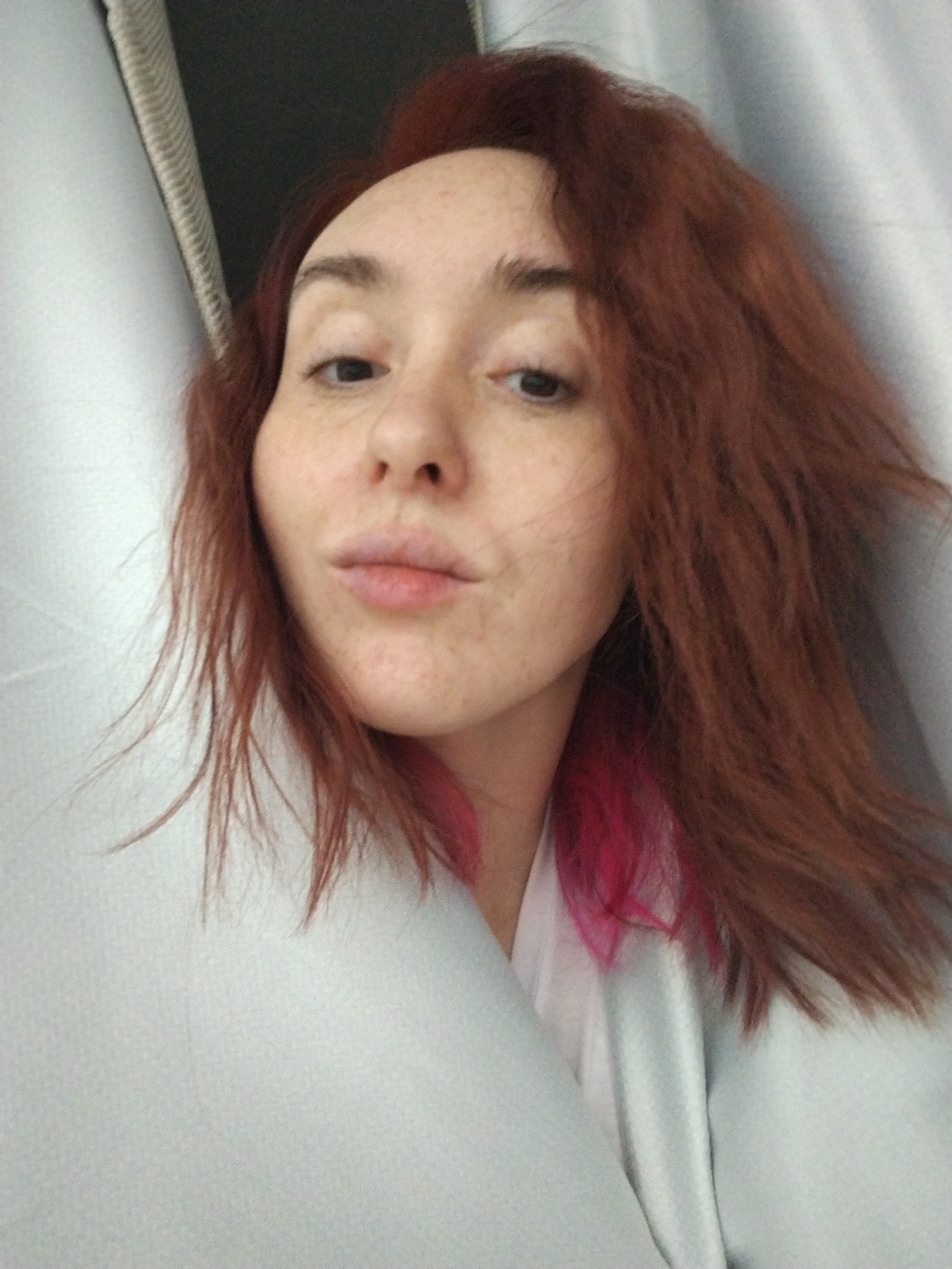 A close-up selfie shows a fair-skinned woman with reddish-brown, slightly tousled hair, with some pink streaks near her neck. She has freckles and is making a slight puckering expression with her lips. She is wearing a light-colored garment that appears to be a robe or a loose shirt. The background consists of neutral-colored curtains or wall coverings. There is no indication in the image to suggest this image was taken in Izhevsk, Russia.