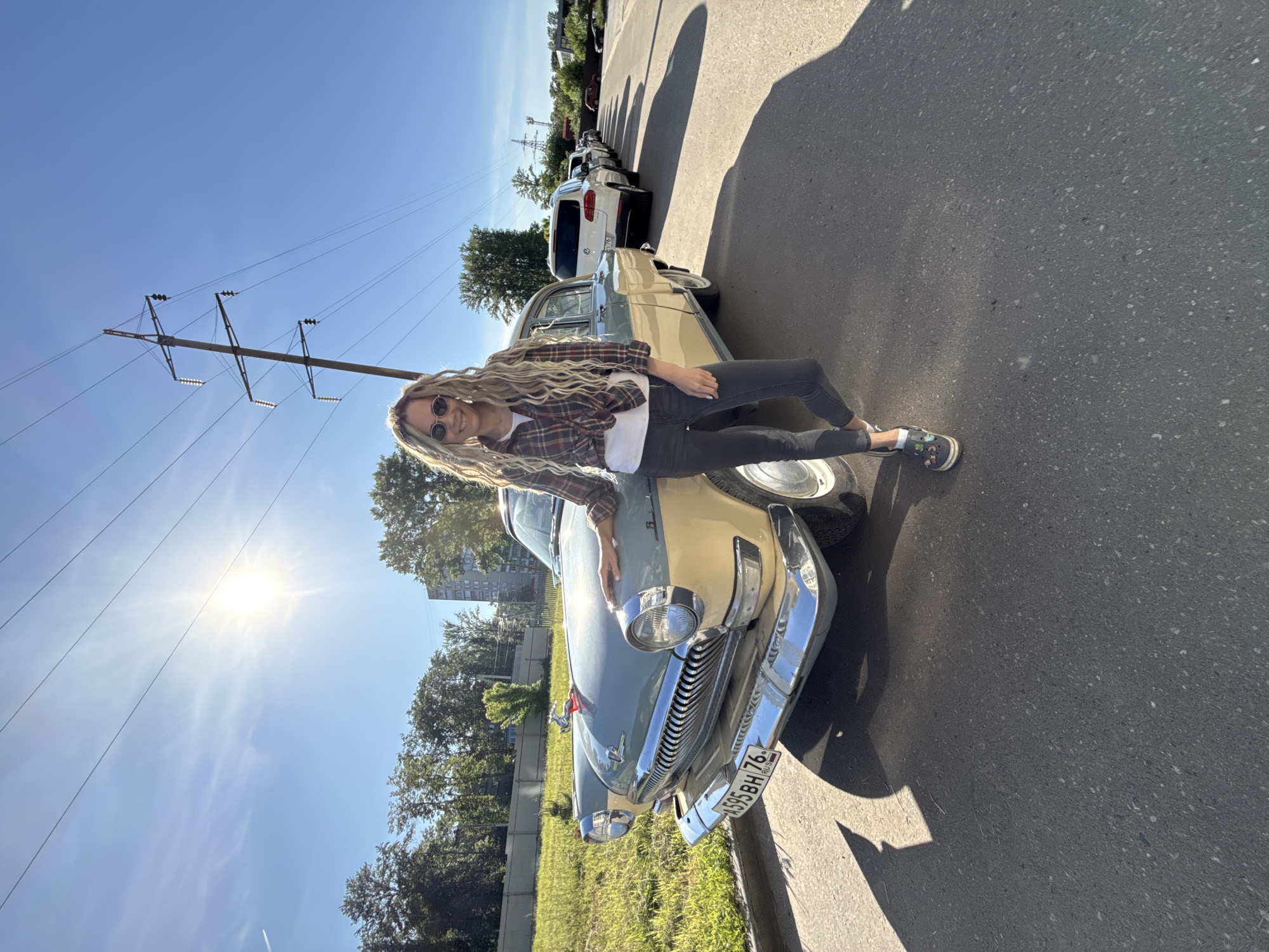 A full-body shot captures a smiling woman posing with a vintage car on a bright, sunny day in what appears to be a roadside setting in Rusanovo, Russia.
The woman has long, wavy blonde hair, wears dark sunglasses, and is dressed in a plaid long-sleeved shirt tied at the waist over a white t-shirt, dark jeans, and black and white sneakers. She is leaning casually on the hood of the car, her left arm resting on it, and her right leg bent at the knee. Her expression is cheerful, with a wide smile directed at the camera.
The car is a classic two-tone model, likely a Soviet-era GAZ-21 Volga, featuring a light blue or teal lower body and a cream or beige upper section. It has prominent chrome accents, including a large grille and bumpers, and a hood ornament. The visible license plate, "X595BH 76 RUS", indicates its registration in the Yaroslavl Oblast region of Russia.
The setting is outdoors, alongside a paved road with a curb and a grassy verge. In the background, a line of modern cars is parked along the street, and green trees and some buildings are visible further in the distance. The sky is clear and bright blue, dominated by the intense sun which causes a noticeable lens flare. A large power line pole with multiple cross-arms and wires is also visible against the sky. The strong sunlight creates distinct shadows on the road and the car. The overall impression is one of a relaxed, sunny day.